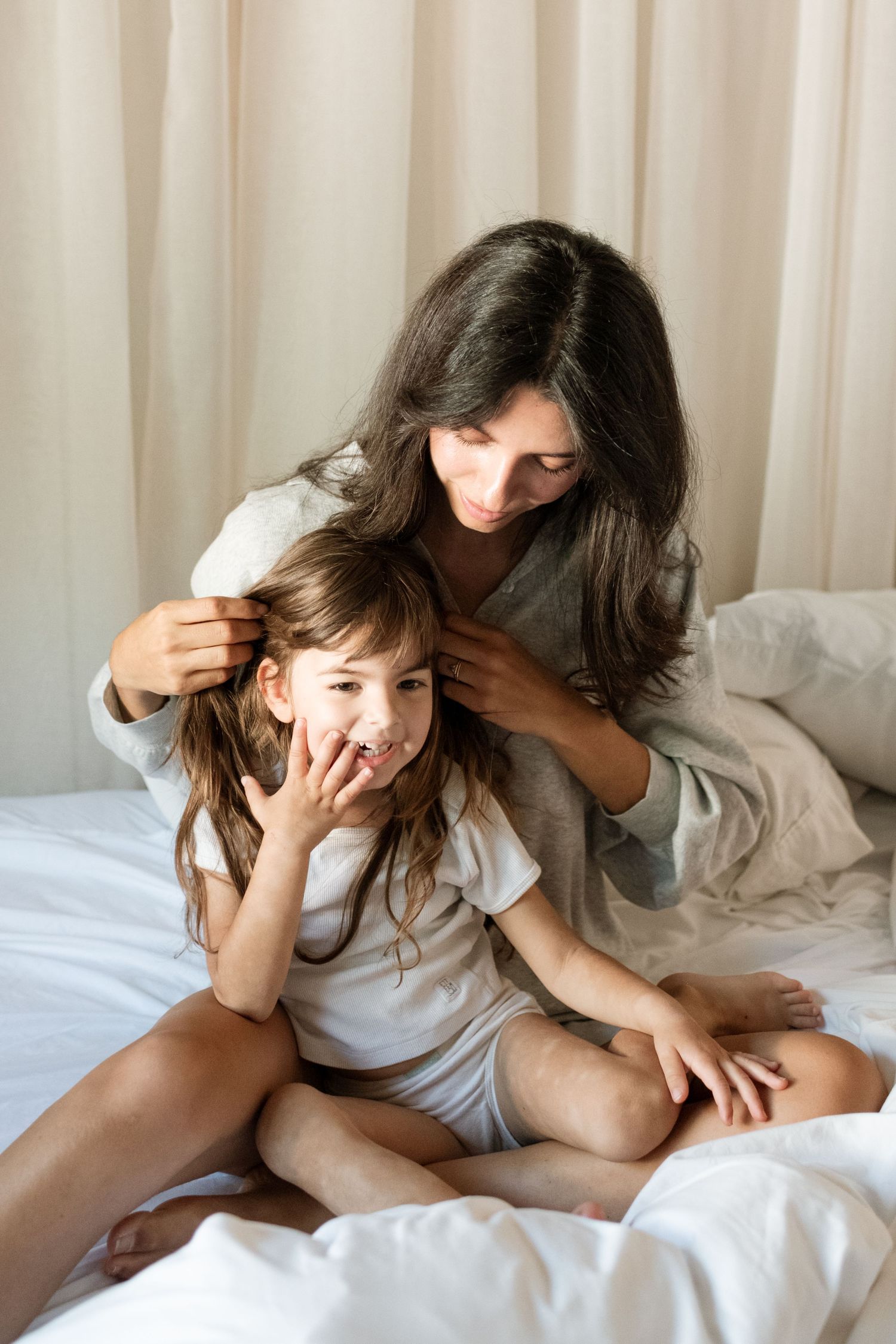 A mother with dark hair sits on a white bed braiding her young daughter's hair in matching casual pajamas.
