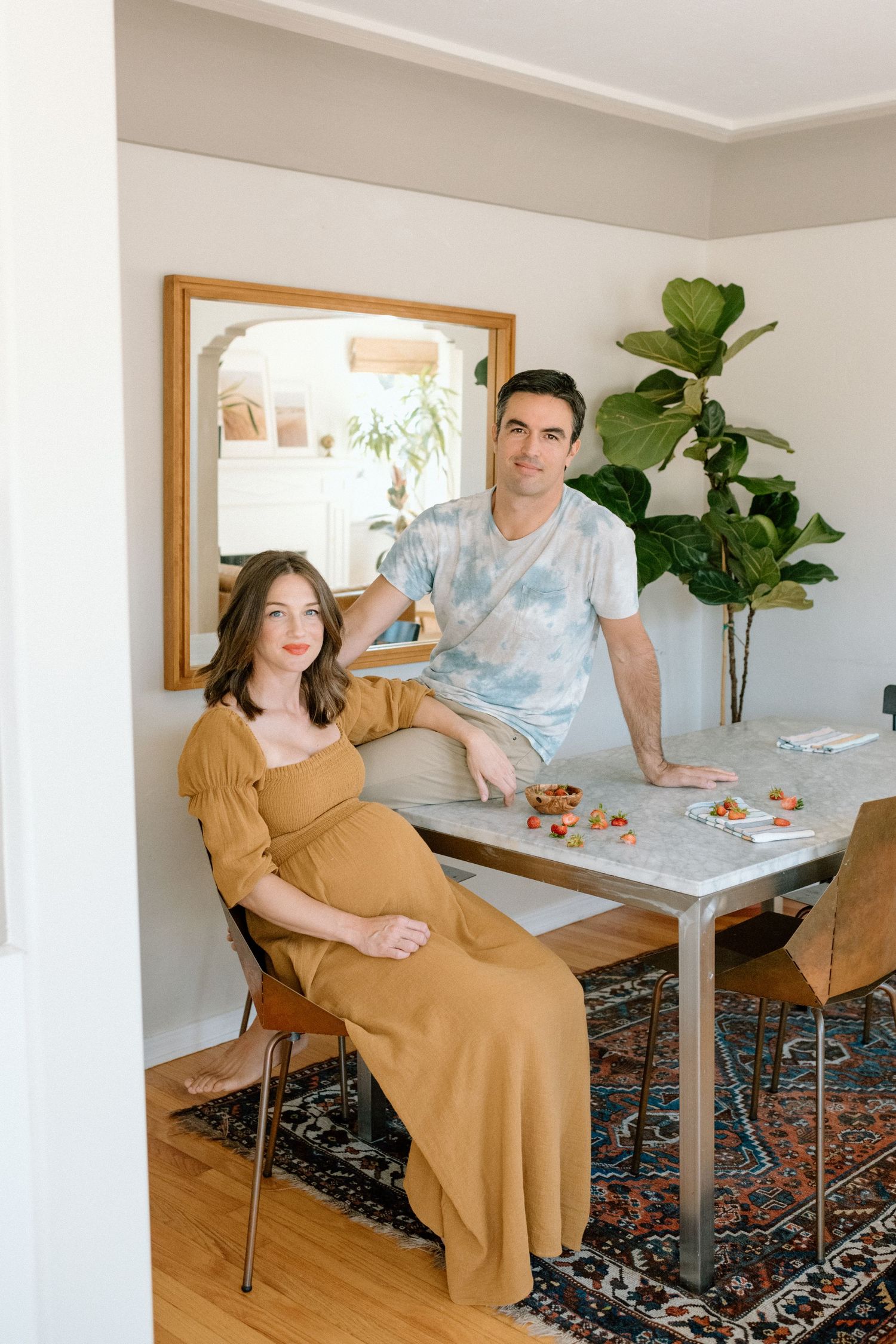 Pregnant woman in golden dress seated at dining table with partner standing beside her in a bright home.