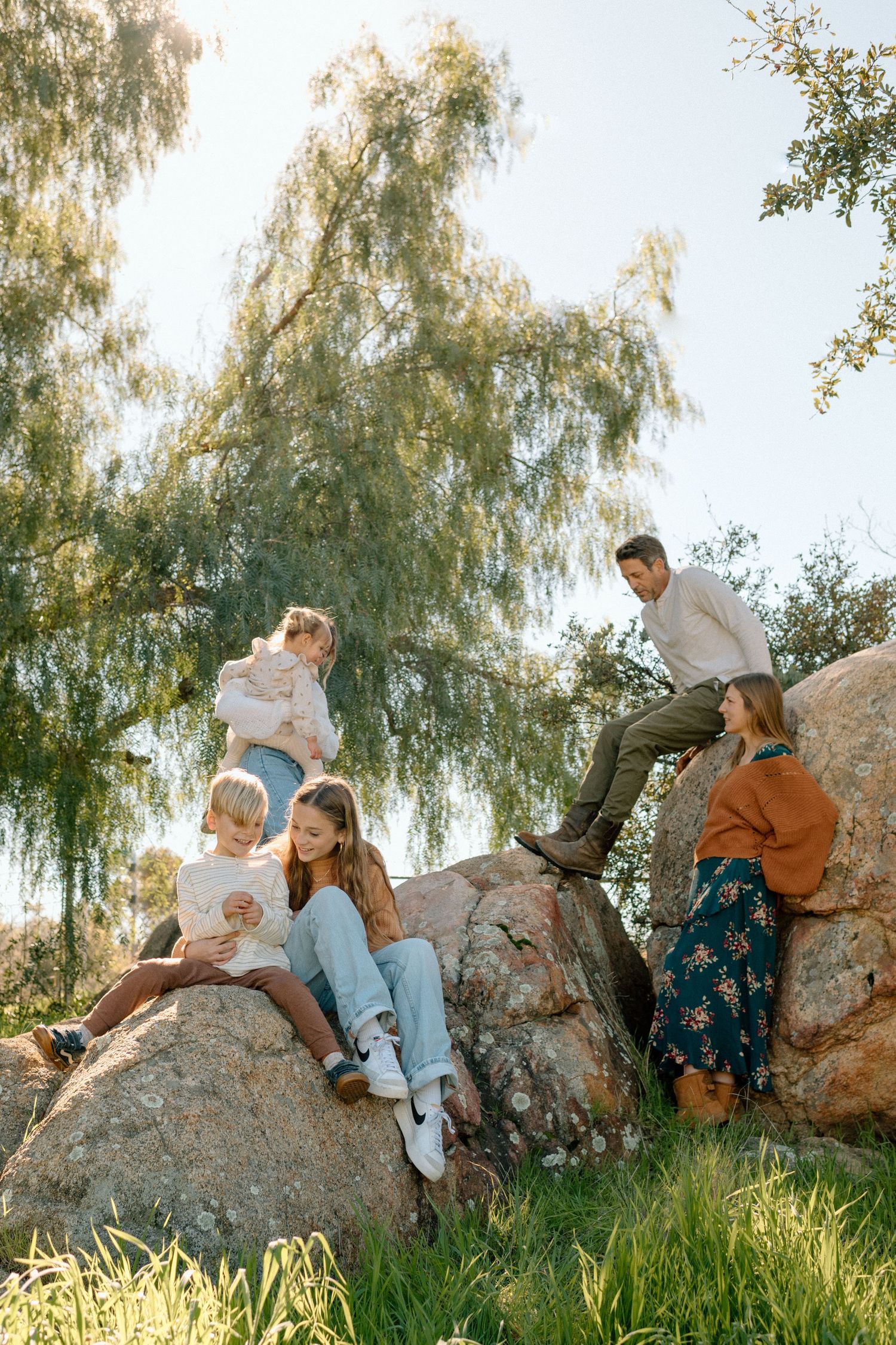 A happy family relaxes on large rocks outdoors, surrounded by lush green trees in warm sunlight.