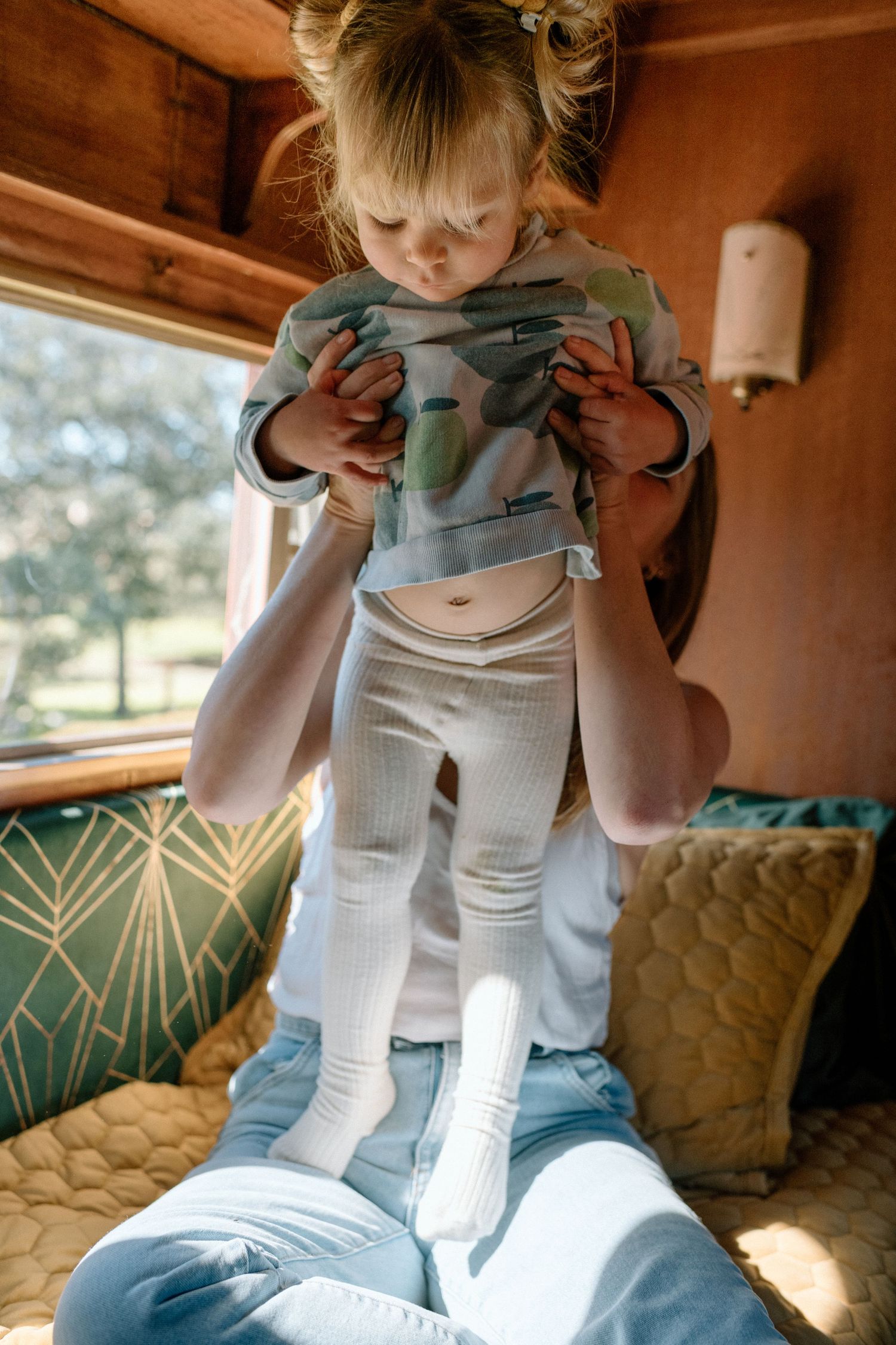 A mother lifts her toddler daughter playfully inside a cozy camper van with warm wood interiors.