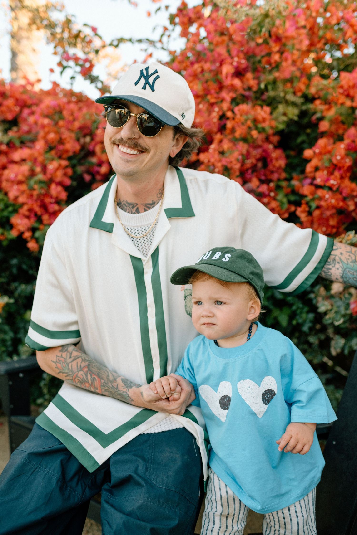 Tattooed father in NY Yankees cap poses with his toddler in a Cubs hat amid red flowers.