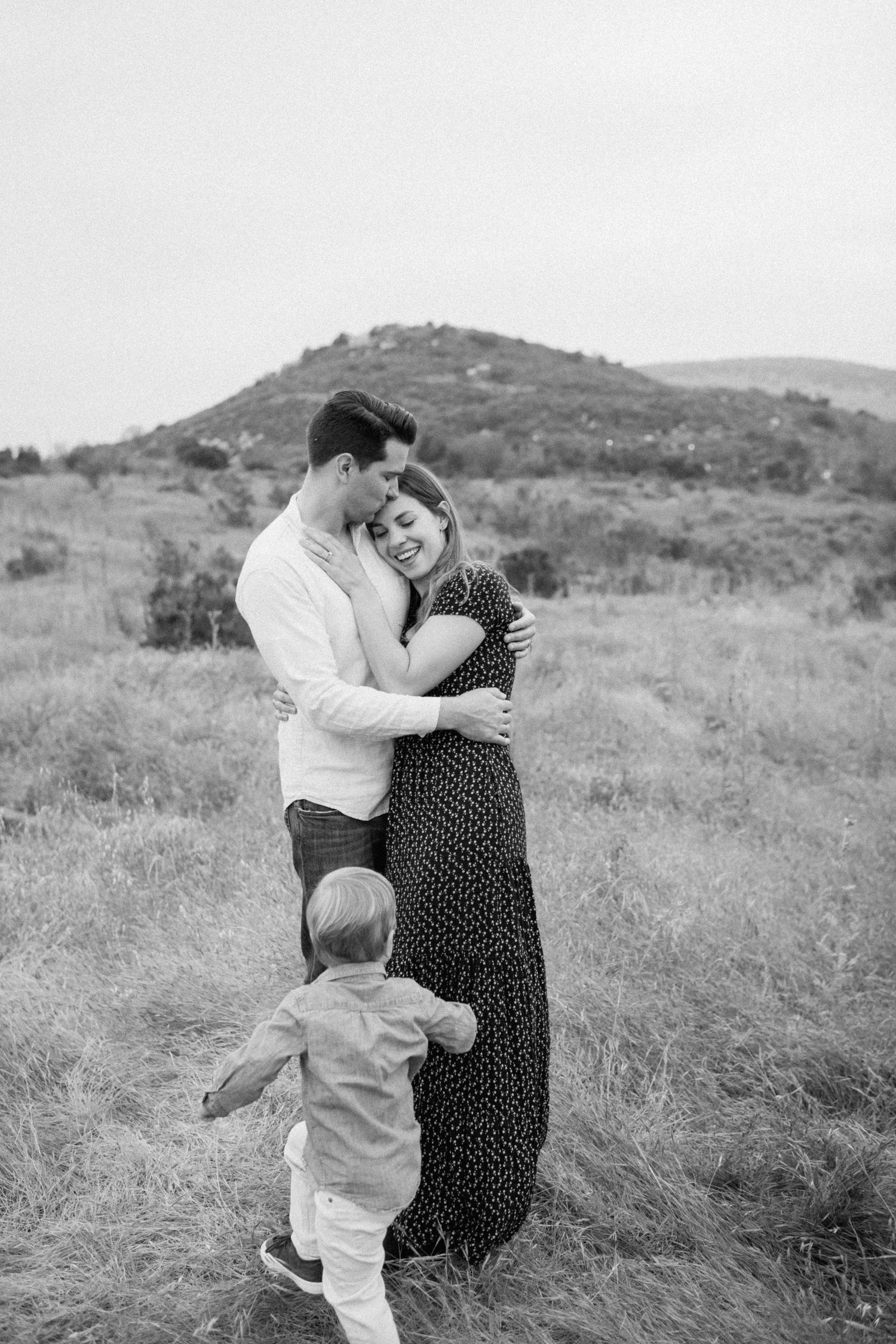 Couple embracing and laughing in an open field while toddler reaches up to them, black and white.