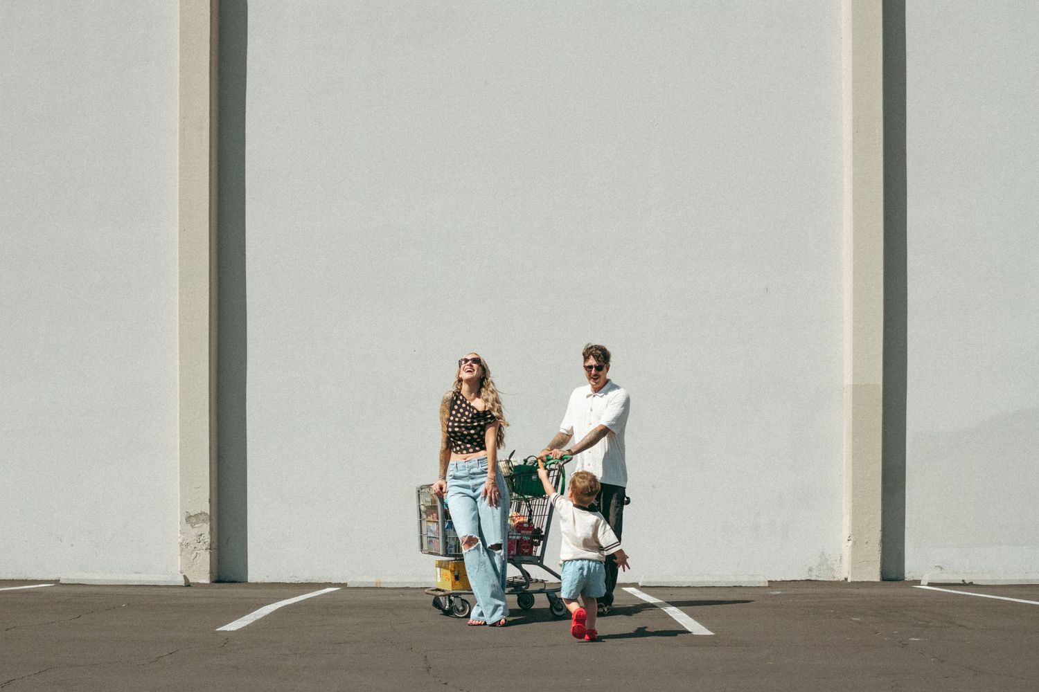 Two people standing in a parking lot with shopping bags and a cart against a white wall.