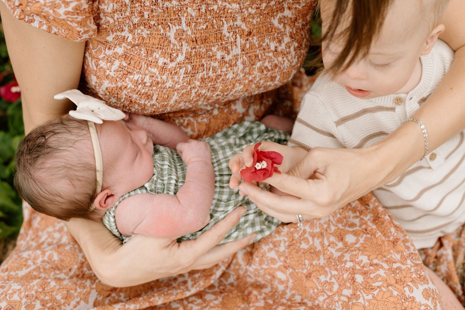 Mother holds newborn baby while young child offers a small red flower during a family photo session outdoors.
