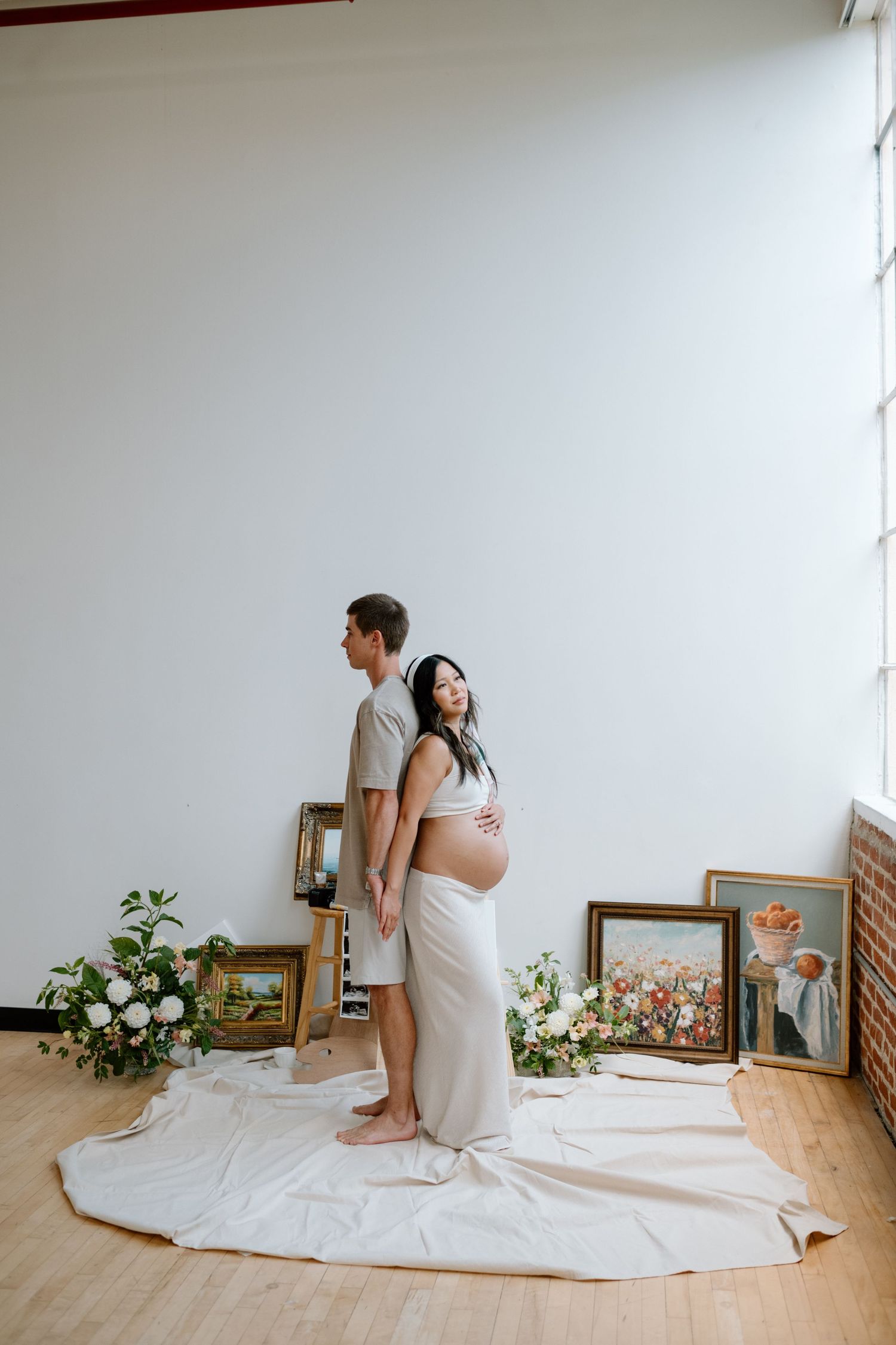 Standing man stands back to back with pregnant wife during maternity photoshoot in a San Diego studio.