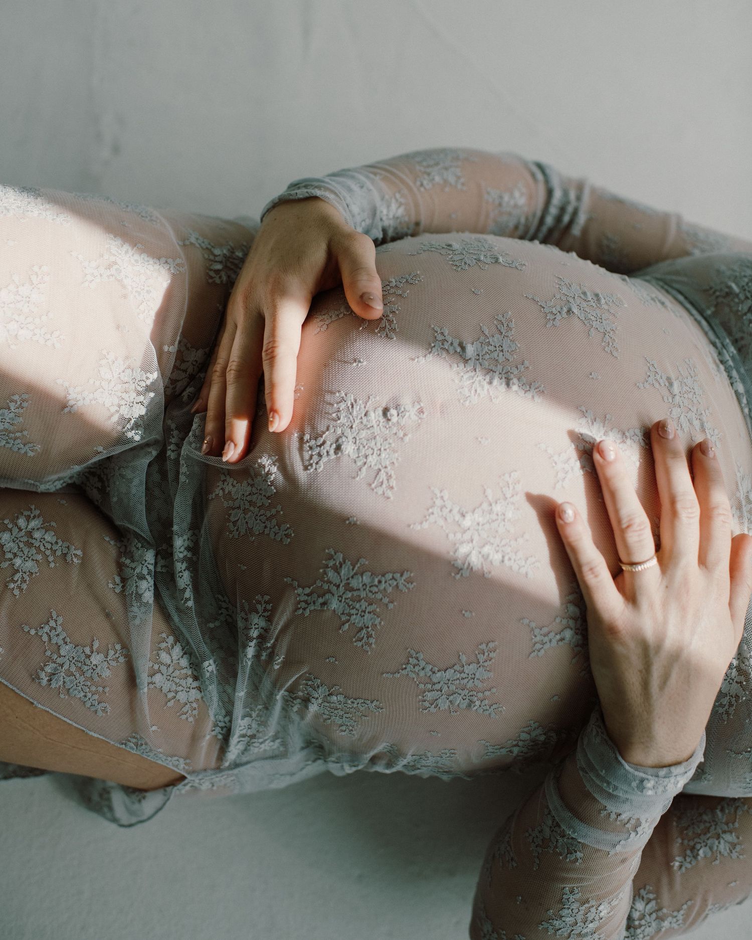Close-up of pregnant woman in floral lace bodysuit gently holding her baby bump with both hands.