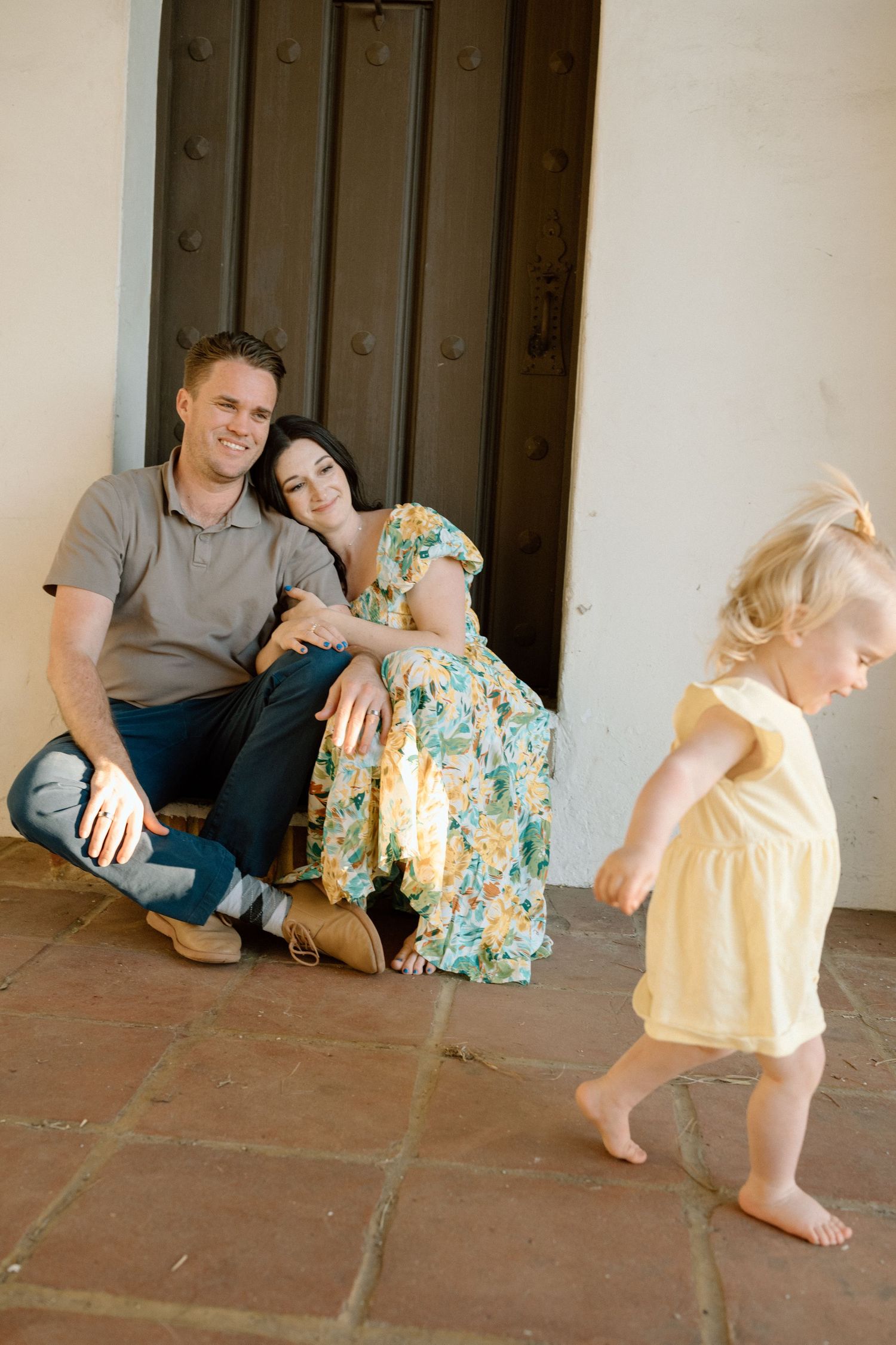 Smiling parents sit against a dark wooden door watching their blonde toddler girl walk barefoot on stone tiles.