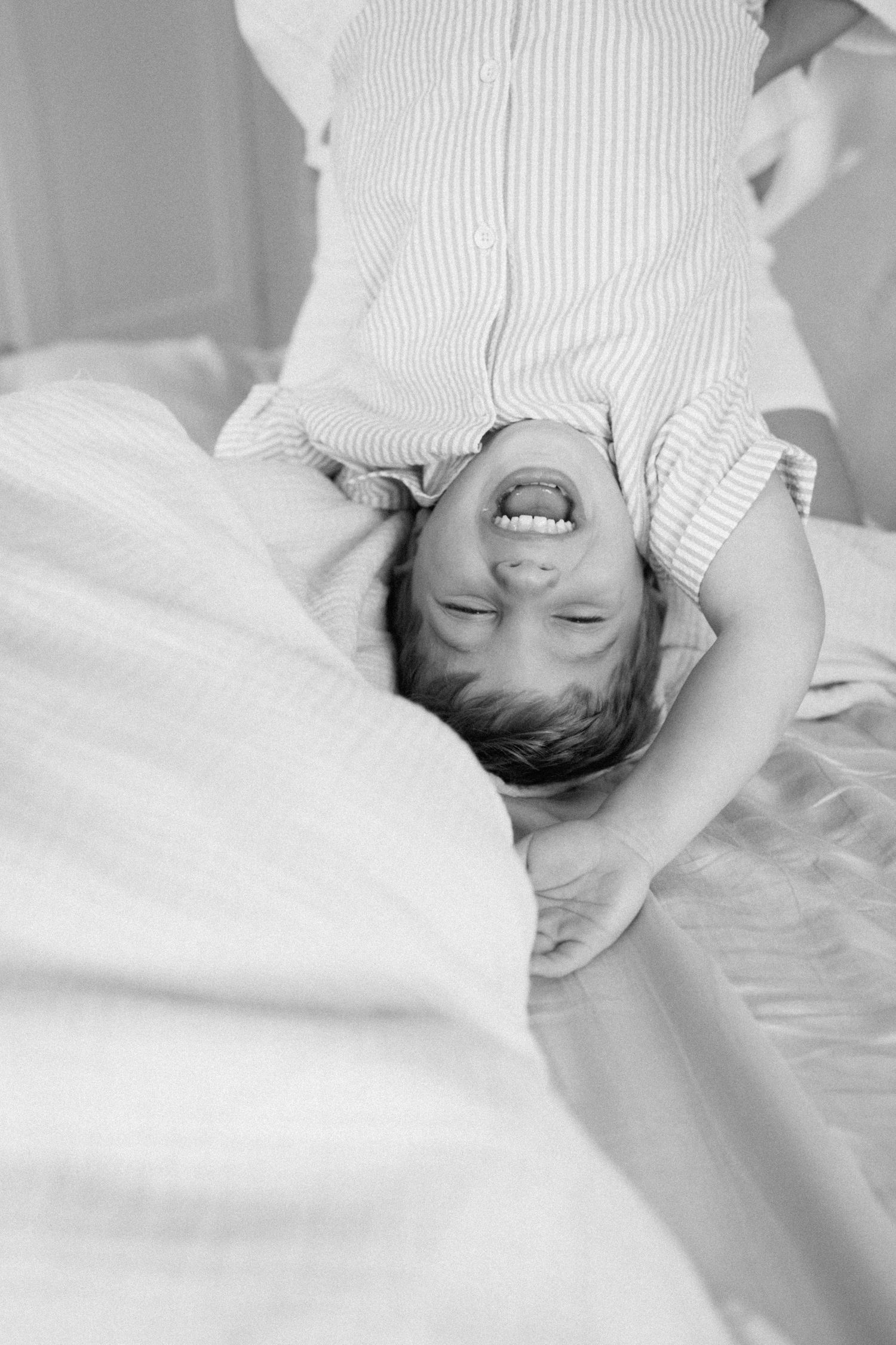 Black and white photo of laughing child lying upside down on a white bed.
