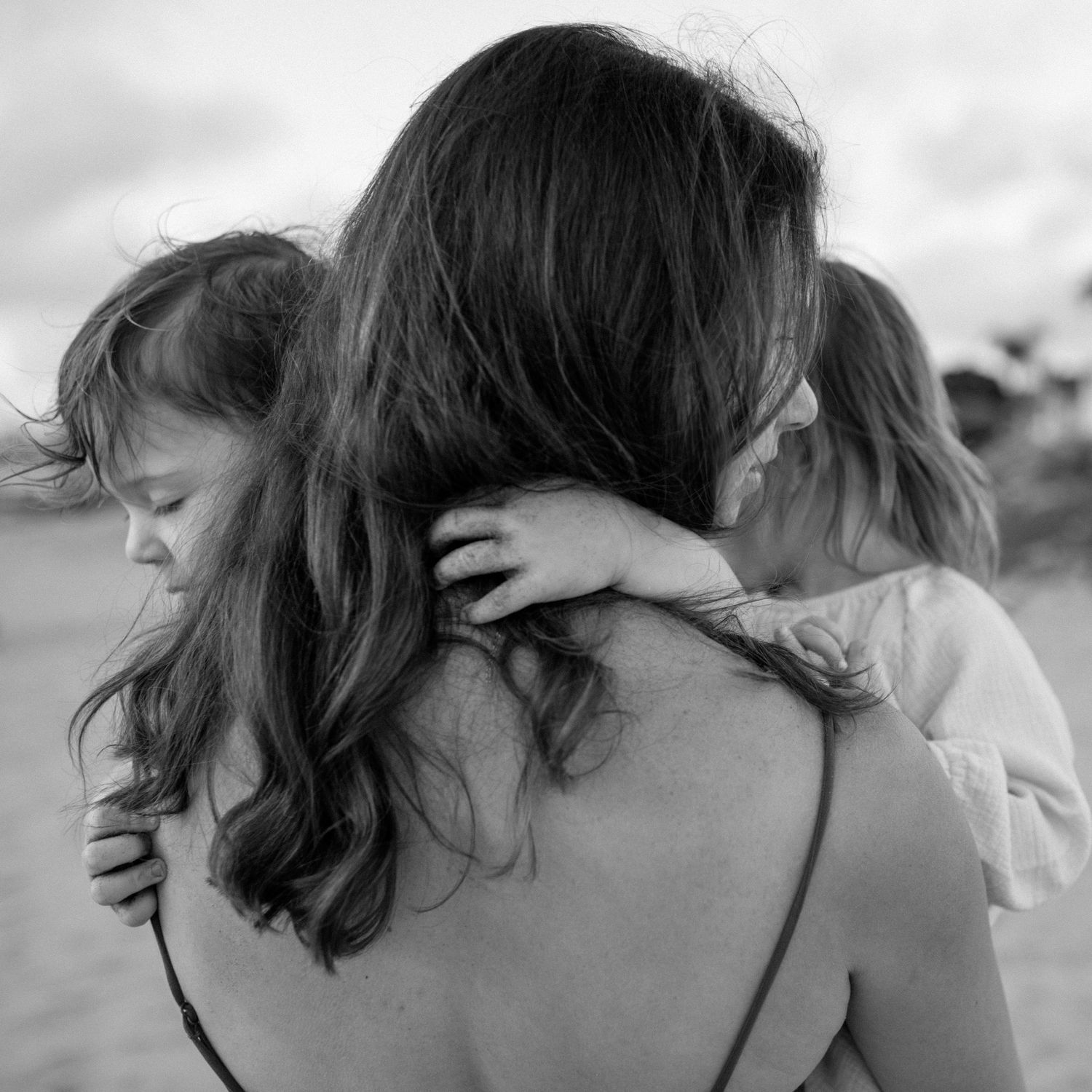 Black and white image of a mother carrying two young daughters on her back at the beach.
