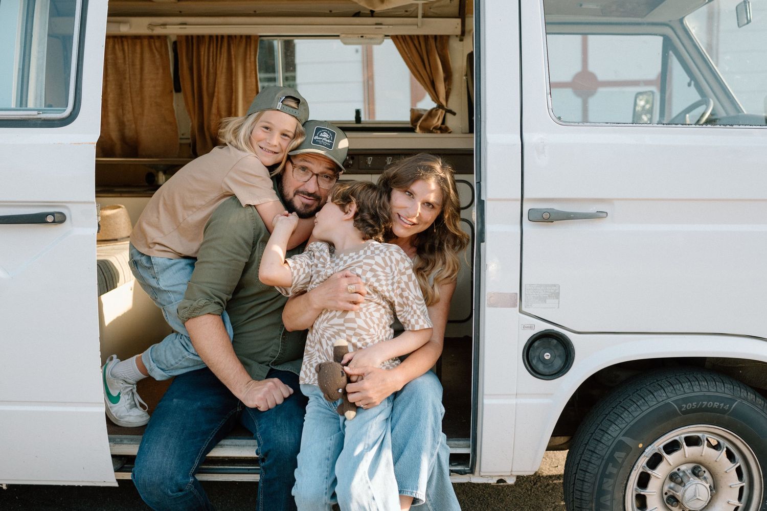 Happy family of four hugging in the open doors of a vintage white camper van.