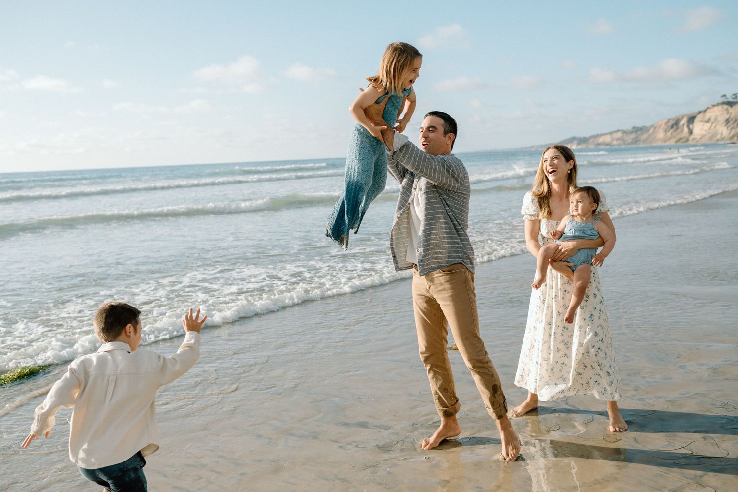 Father lifts daughter joyfully on sunny beach while mother holds baby and son runs nearby.