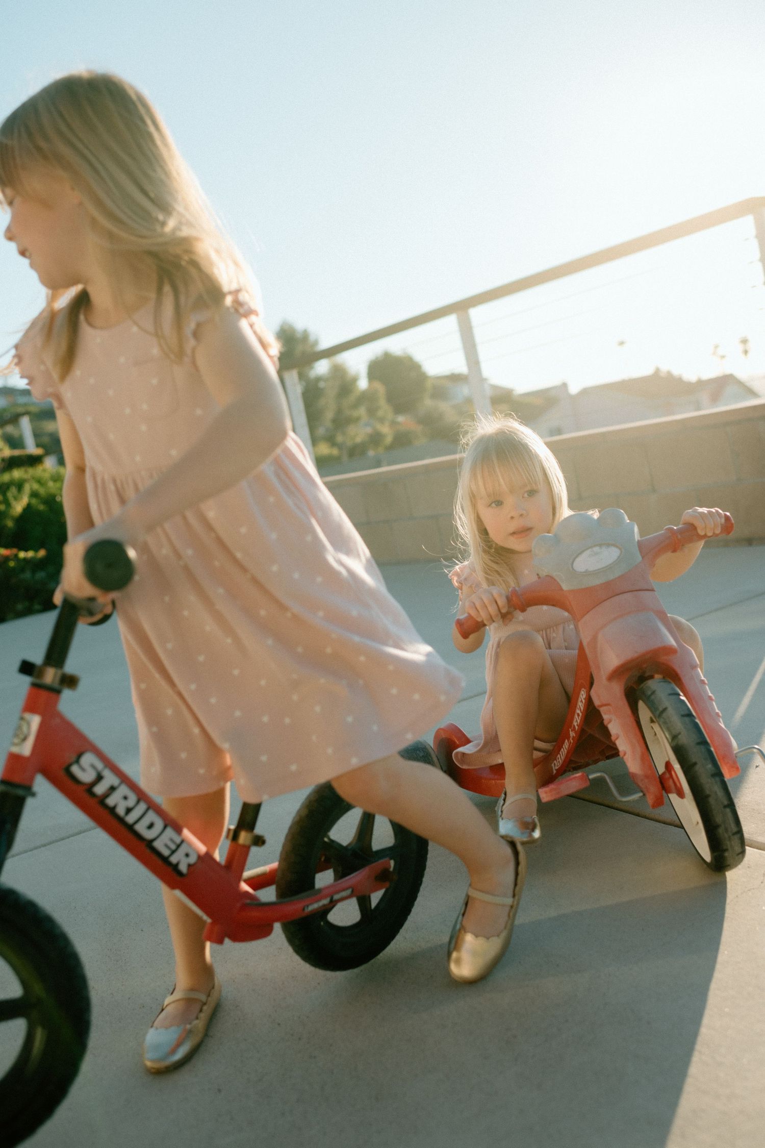 Two young blonde girls riding red Strider balance bikes on sunny outdoor concrete surface.