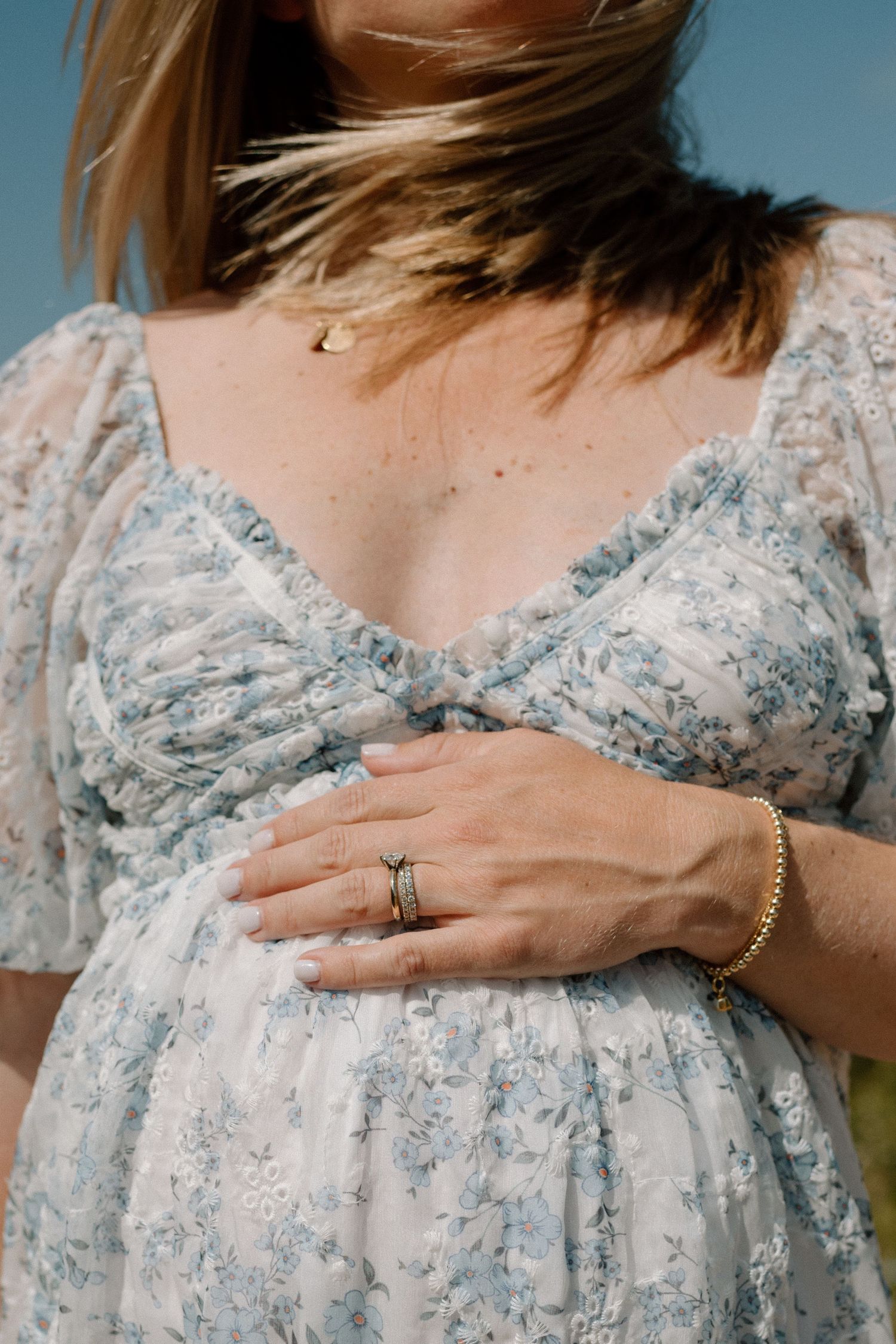 Close-up of pregnant woman in blue floral dress gently cradling baby bump with gold jewelry.