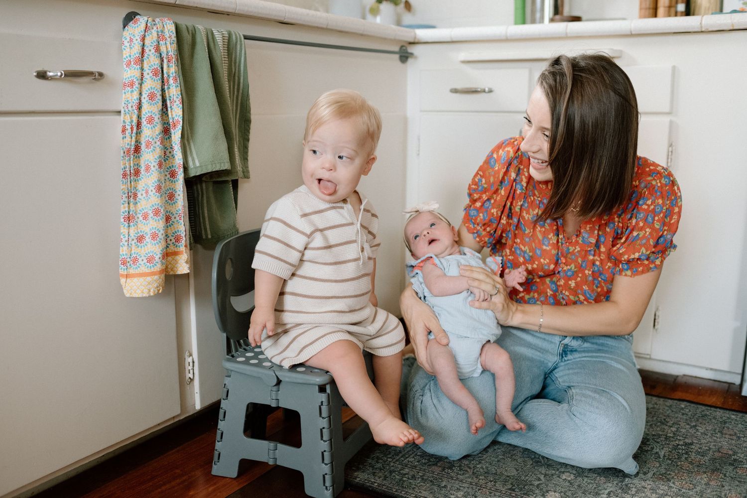 Mother sits on kitchen floor holding newborn baby while toddler with Down syndrome sits on step stool beside her.