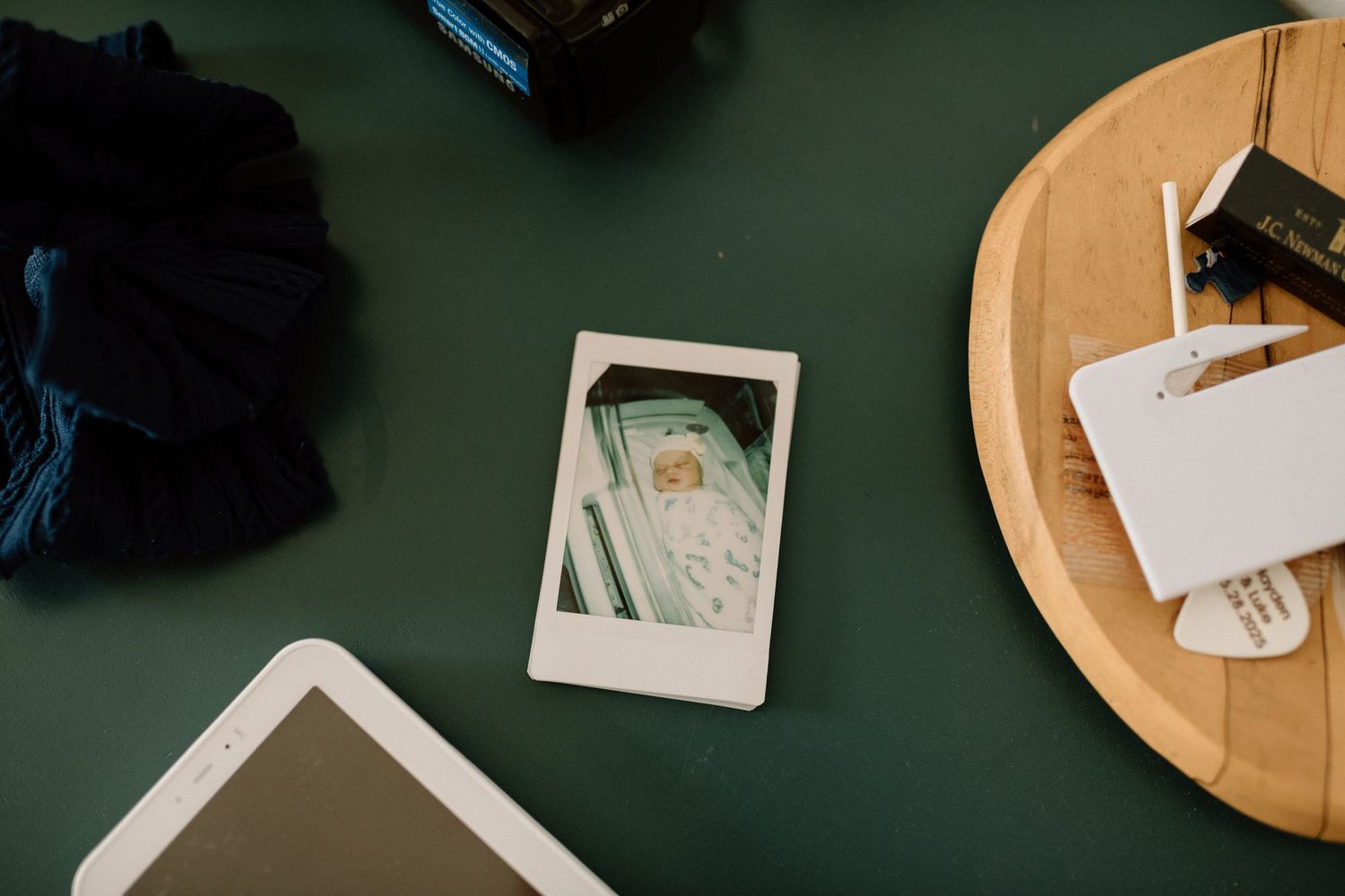 Flat lay of a Polaroid photo, tablet, wooden board, and accessories on a dark green surface.