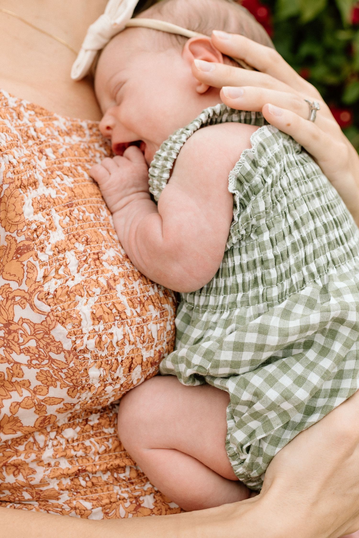 Newborn baby in green gingham romper sleeping peacefully in mother's arms wearing floral dress.