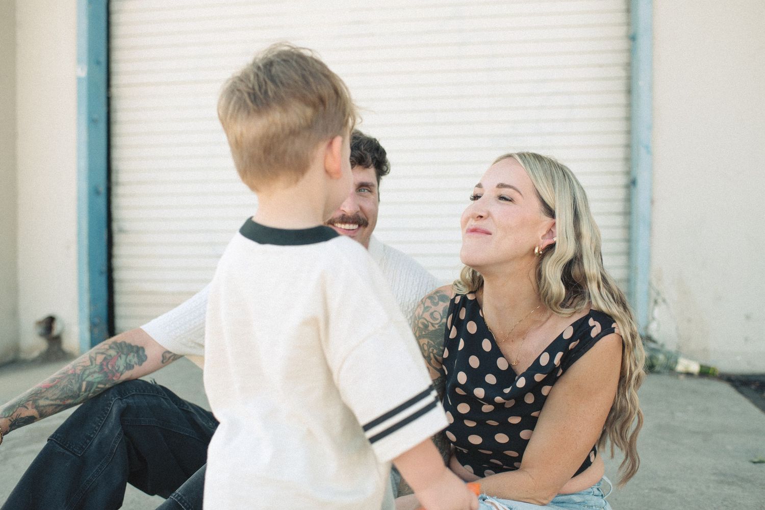 A woman and man smiling at a young blond boy outdoors near a white garage door.