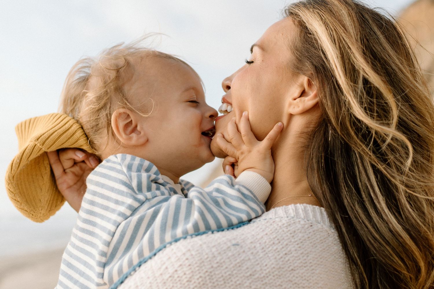 A mother holds her baby who gives her a sweet kiss on the cheek outdoors on a sunny day.