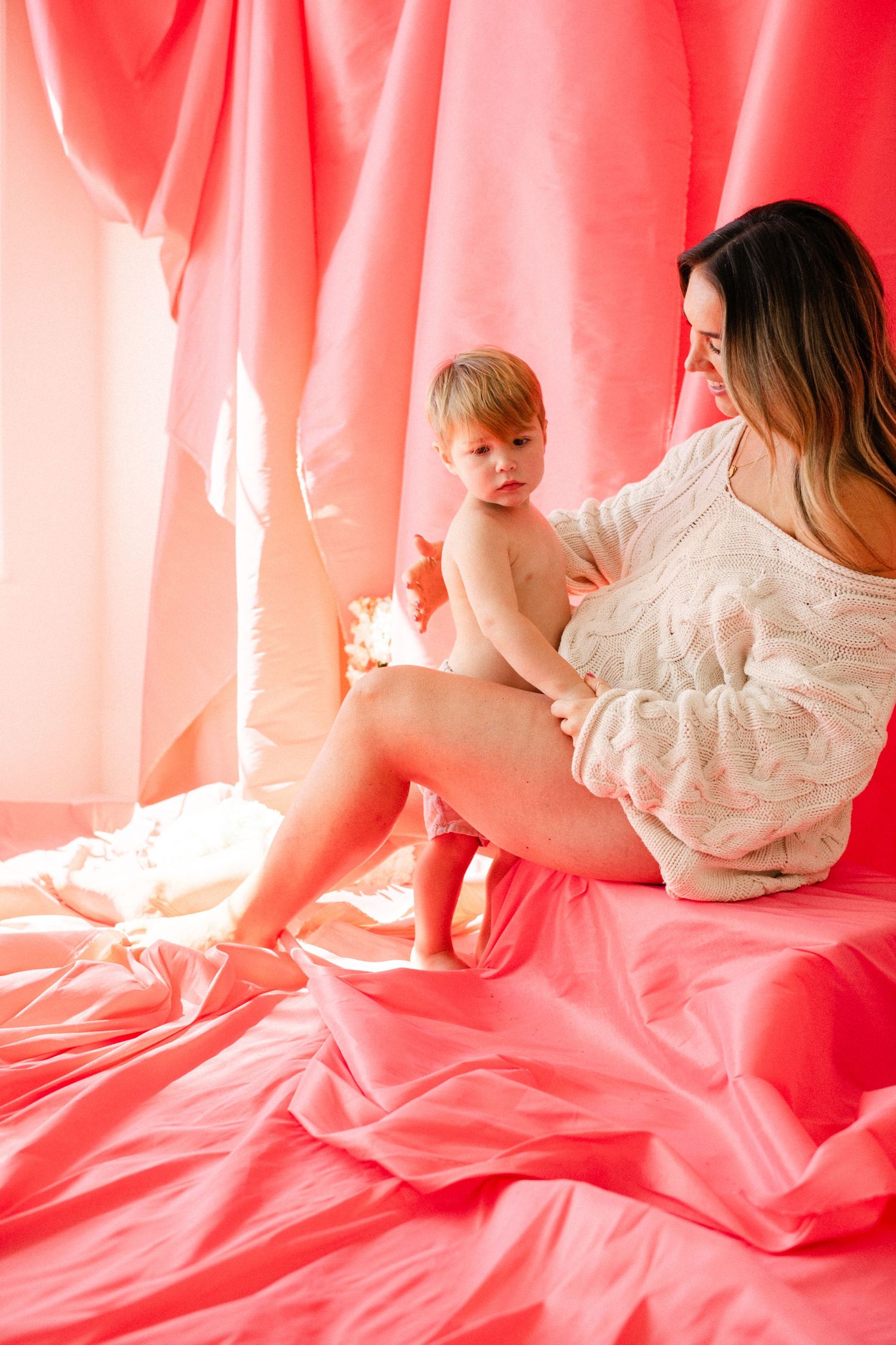 A mother sits on pink bedding holding her toddler, surrounded by soft pink curtains in a warm studio setting.