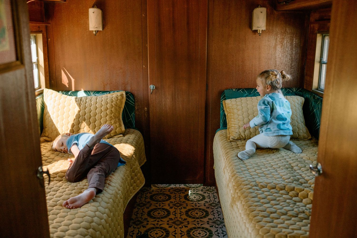 Two children relax on vintage quilted train seats inside a wood-paneled railway compartment.