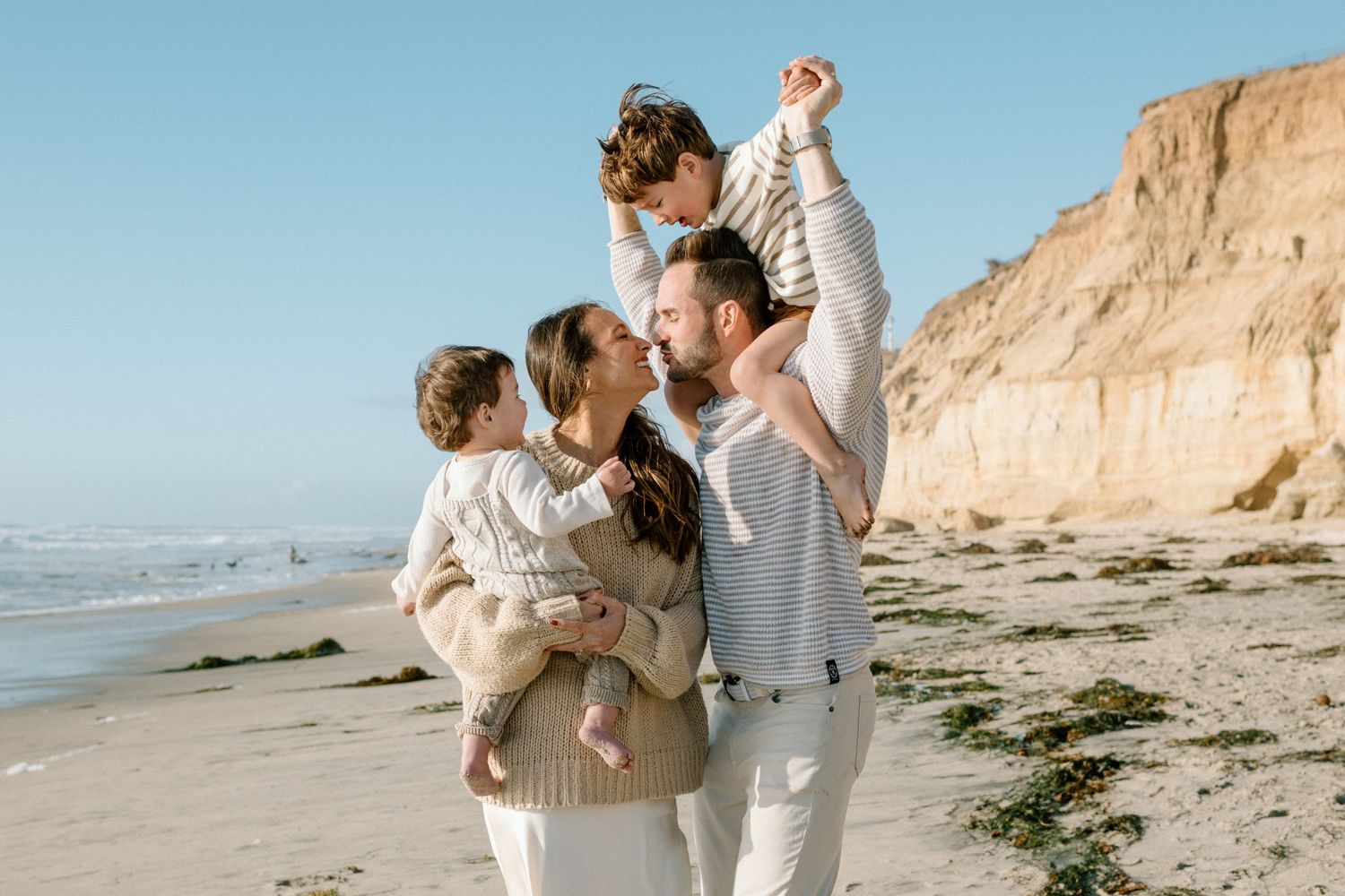 Loving family kissing on beach while father lifts child overhead.