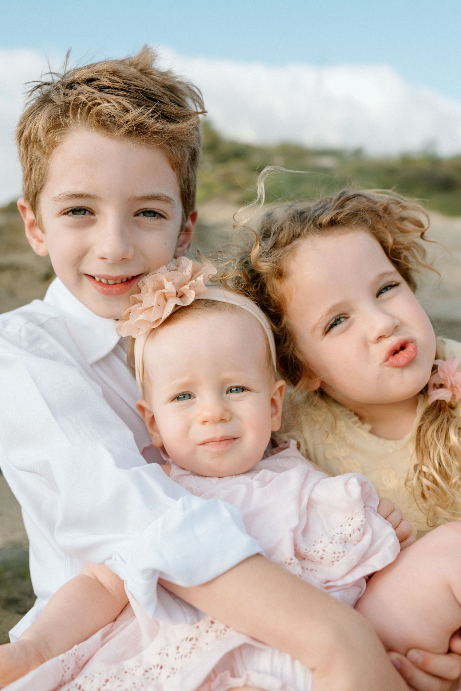 Three siblings posing outdoors, oldest boy and girl hugging baby sister in pink.