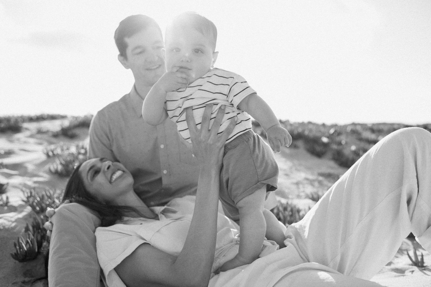 Black and white photo of a happy family with parents and baby playing together on a sunny beach.
