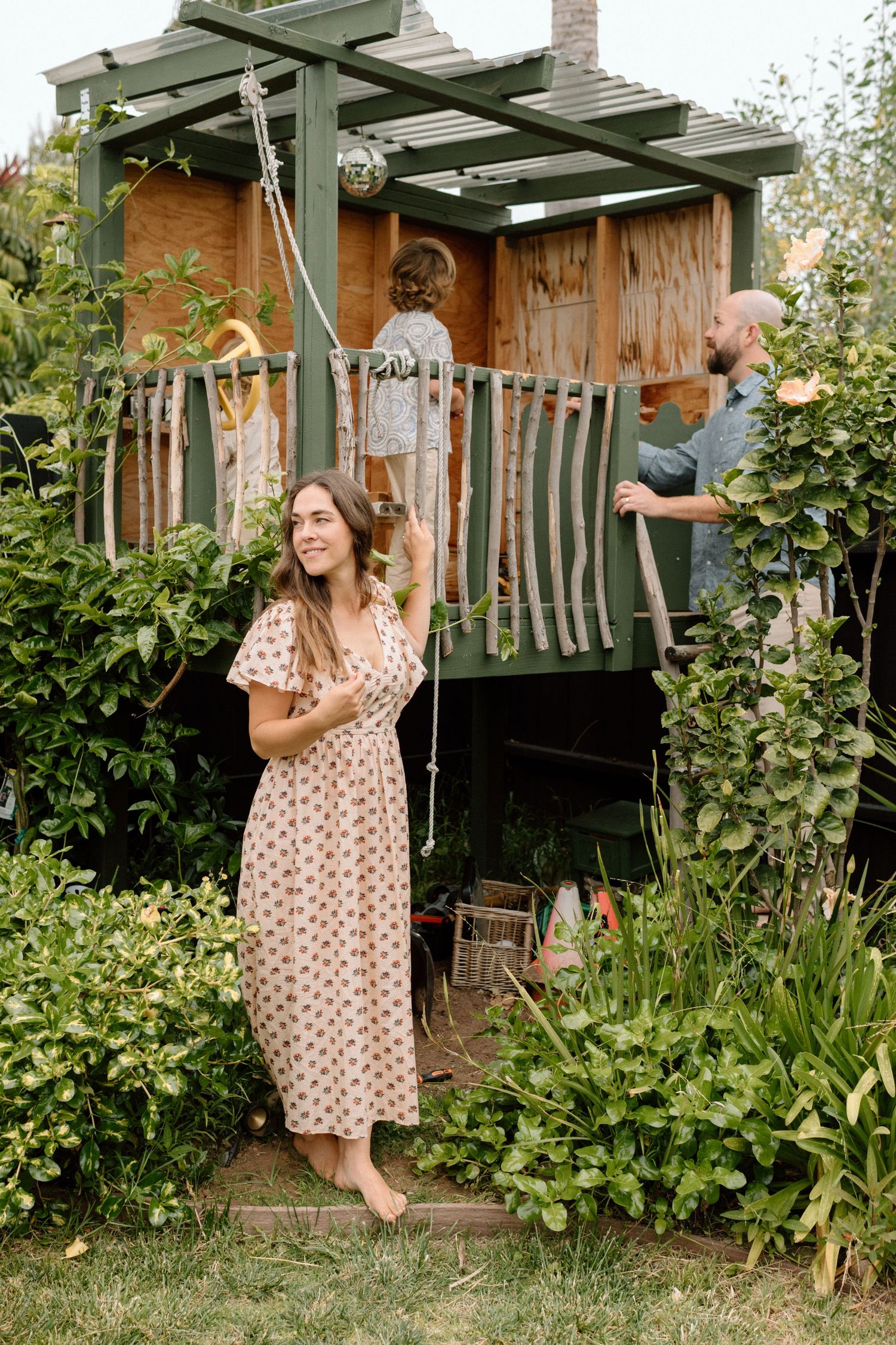 Smiling woman in floral dress stands near a backyard treehouse while man assists child.