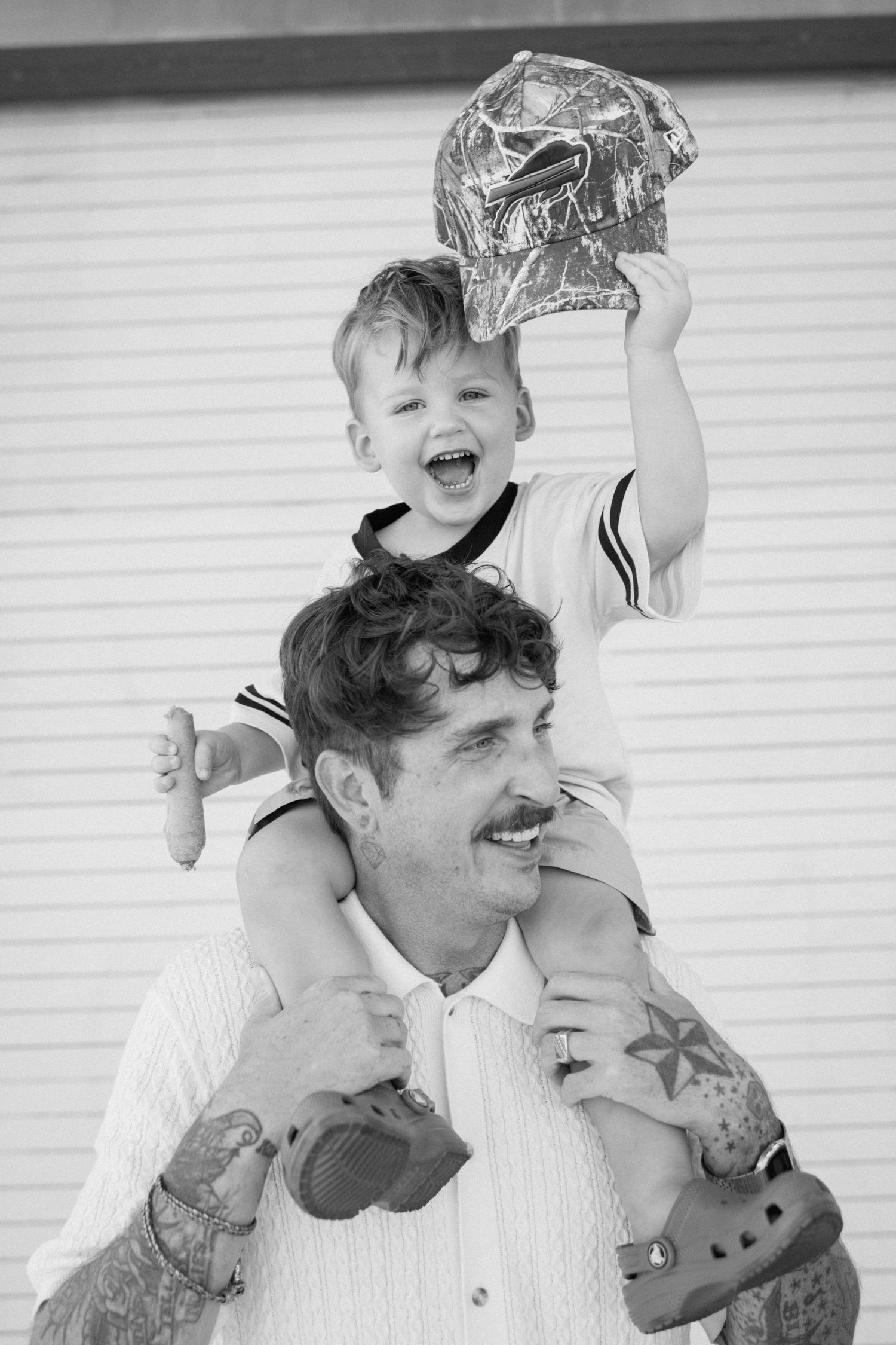Black and white photo of a laughing toddler sitting on a tattooed man's shoulders, holding up a camo cap.