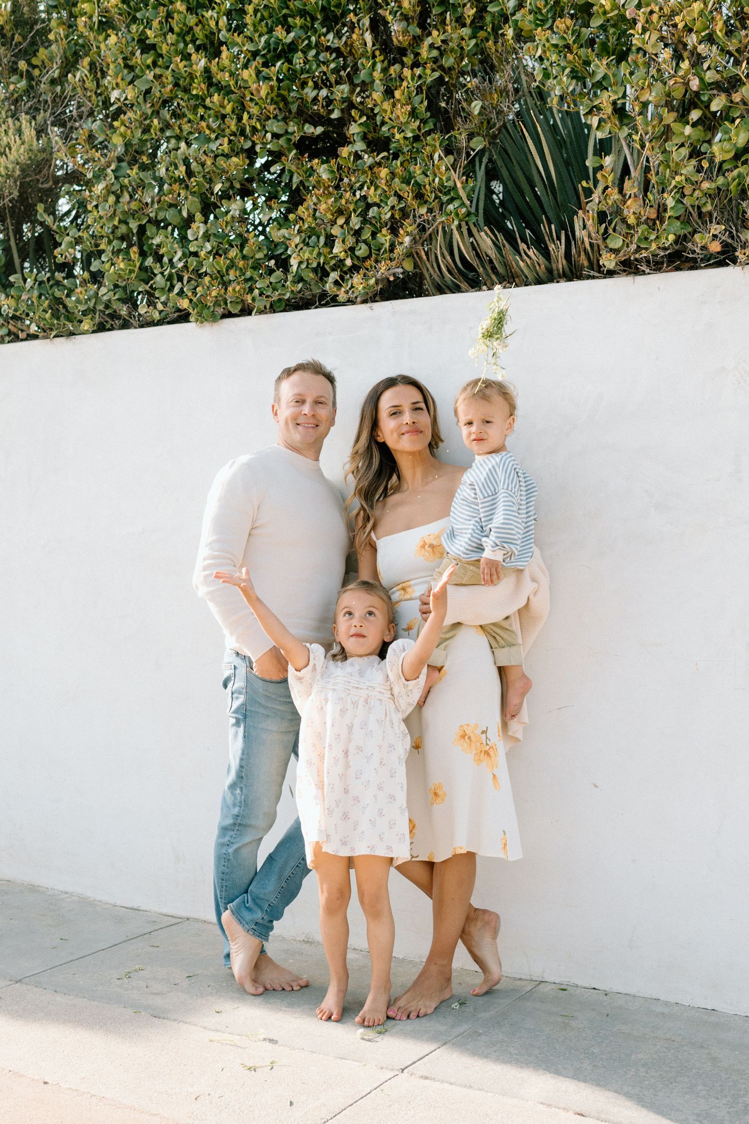 Family of four posing against white wall outdoors, parents holding children in light summer clothing.