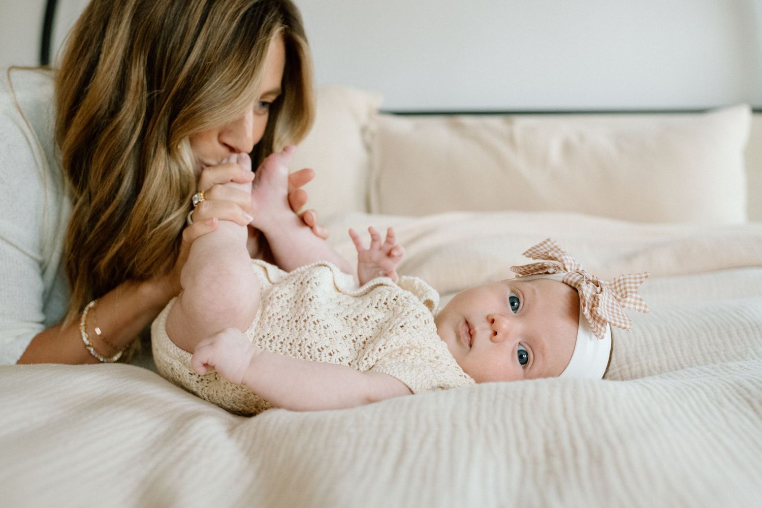 A mother tenderly kisses her newborn baby's hand while the infant lies on a white bed wearing a crochet outfit.