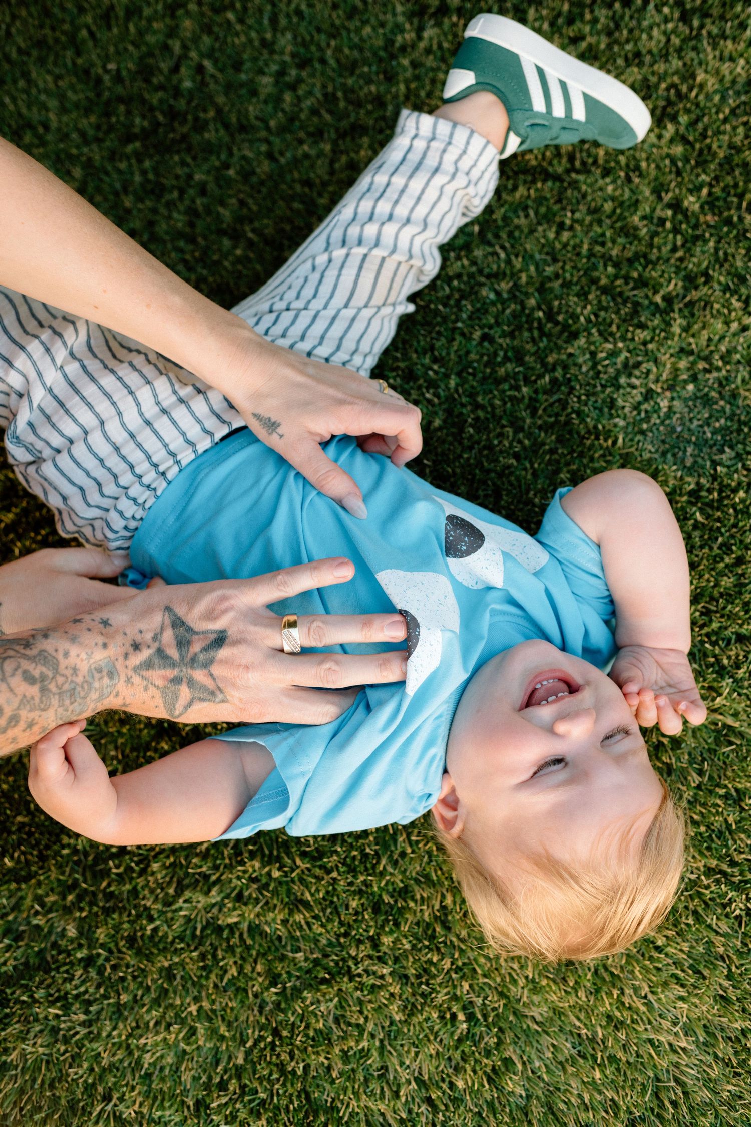 A toddler in blue romper and striped pants lies on green grass with an adults hand showing a tattoo.