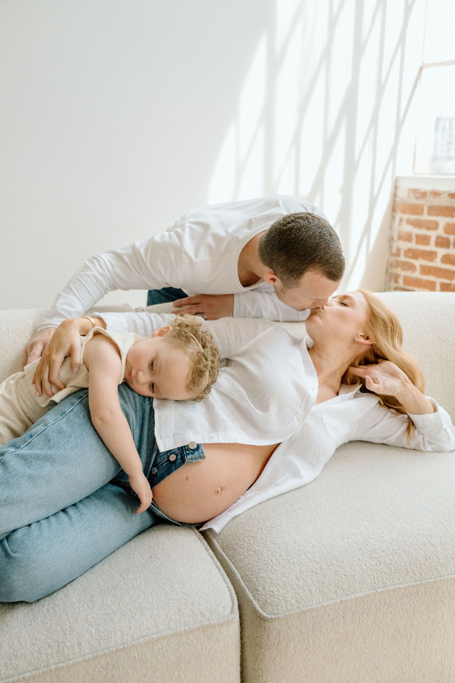 Pregnant woman lying on sofa kissing husband while toddler rests on her belly in bright living room.