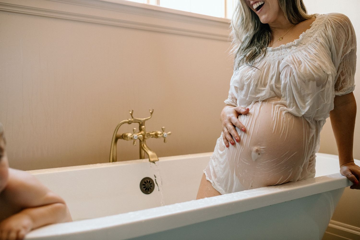 Smiling pregnant woman in white lace dress cradling baby bump while standing beside a vintage white clawfoot bathtub.