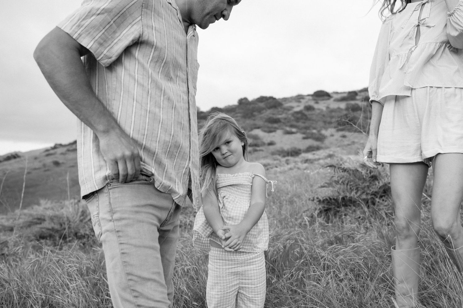 A young girl smiles shyly while holding hands with her parents in a grassy field.