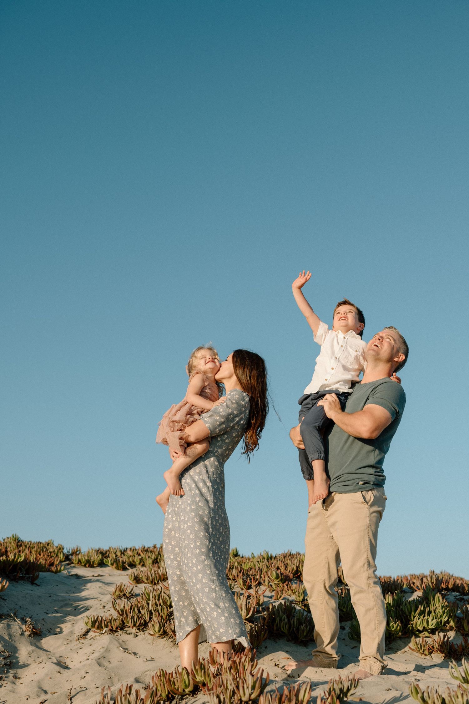 Happy family of four enjoying sunny day outdoors, parents holding young children up high.