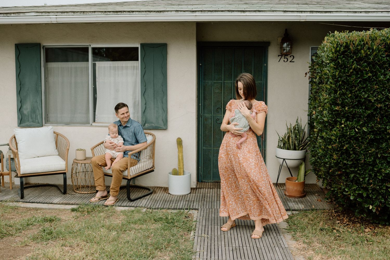 Couple smiling outside a cozy home, woman in floral dress holding a vase, man seated on patio furniture.
