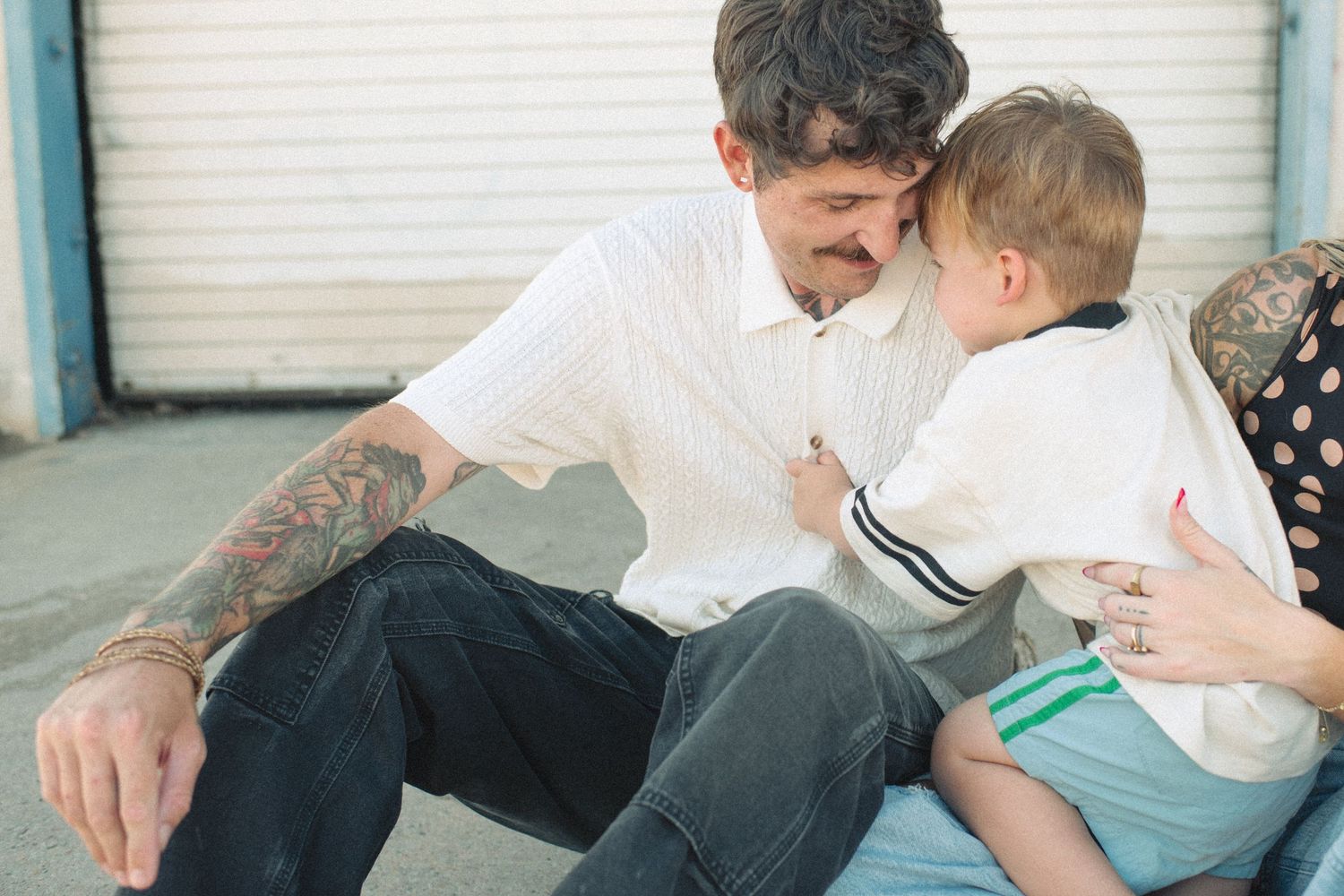 Tattooed father sitting outdoors bonding with his young toddler son in matching white shirts.
