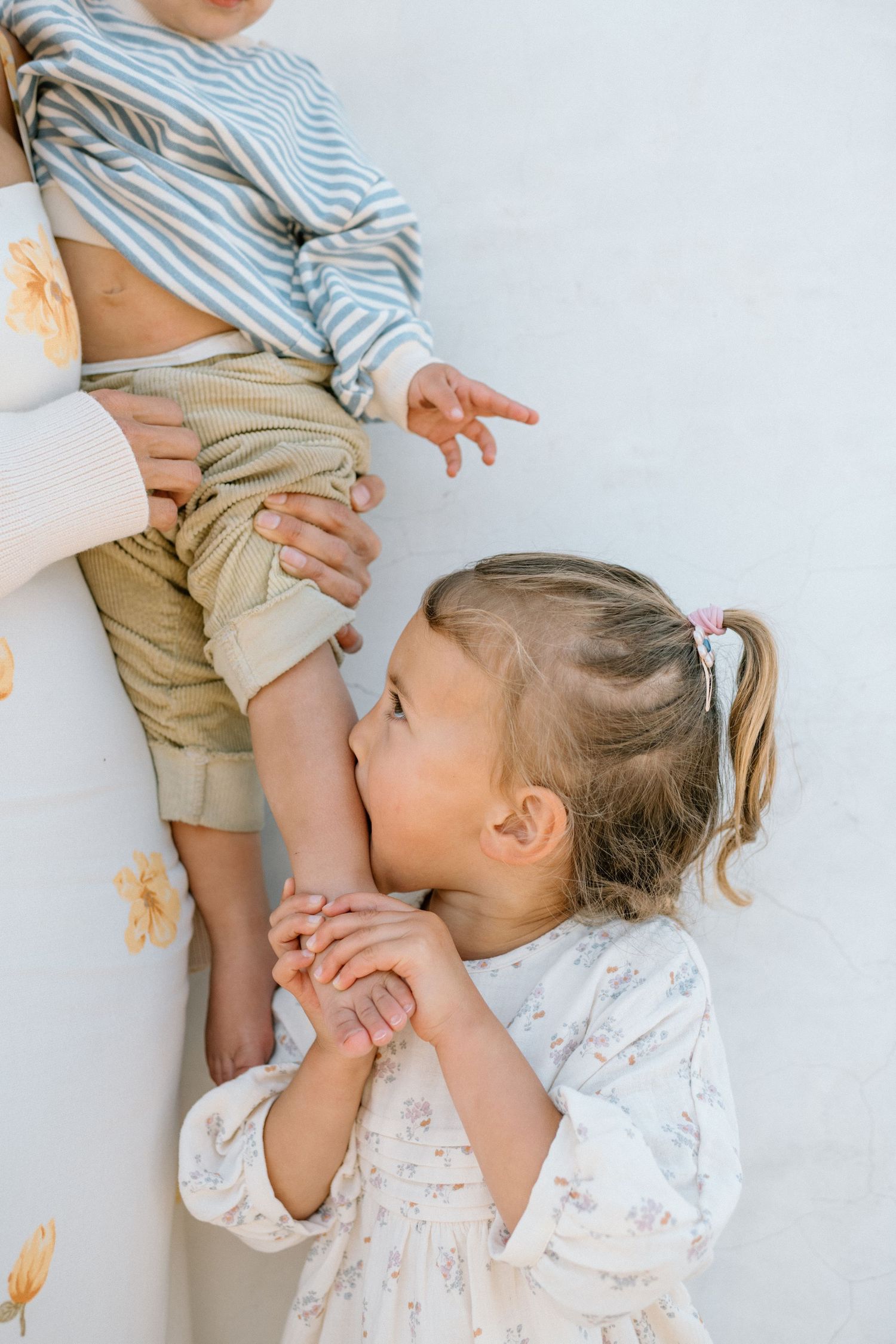 A young girl in a floral dress kisses a toddler's hand while looking up at an adult holding the child.