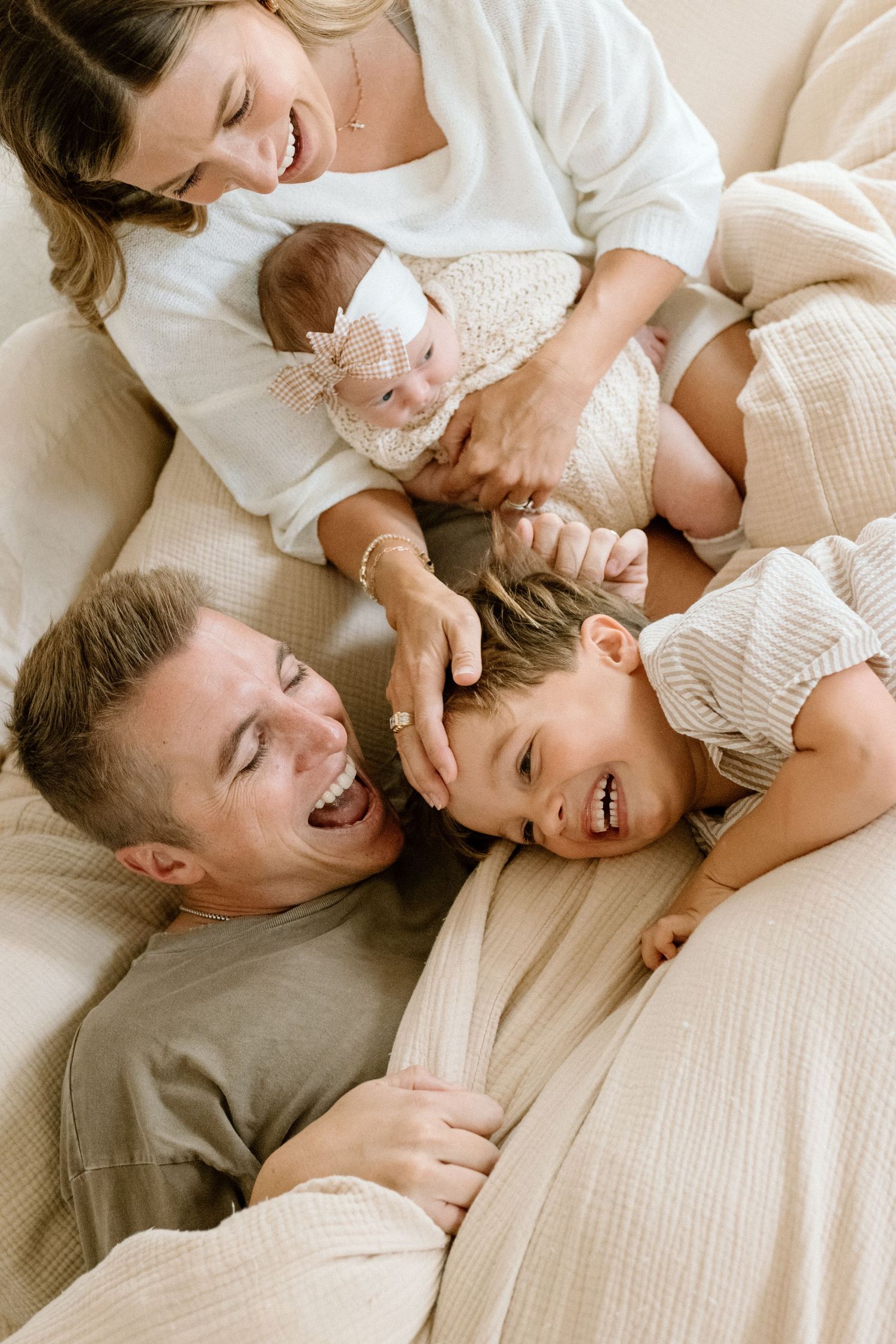 Happy family of four with newborn baby laughing together on a cozy bed in warm neutral tones.