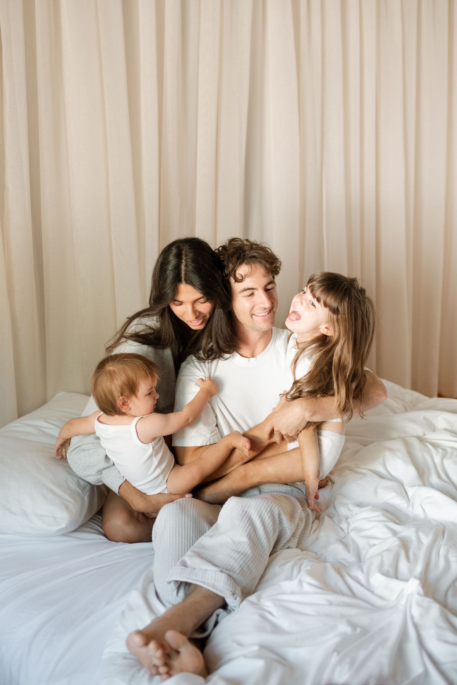 Happy family of four sitting on a white bed, parents smiling at their two young children in a cozy bedroom.