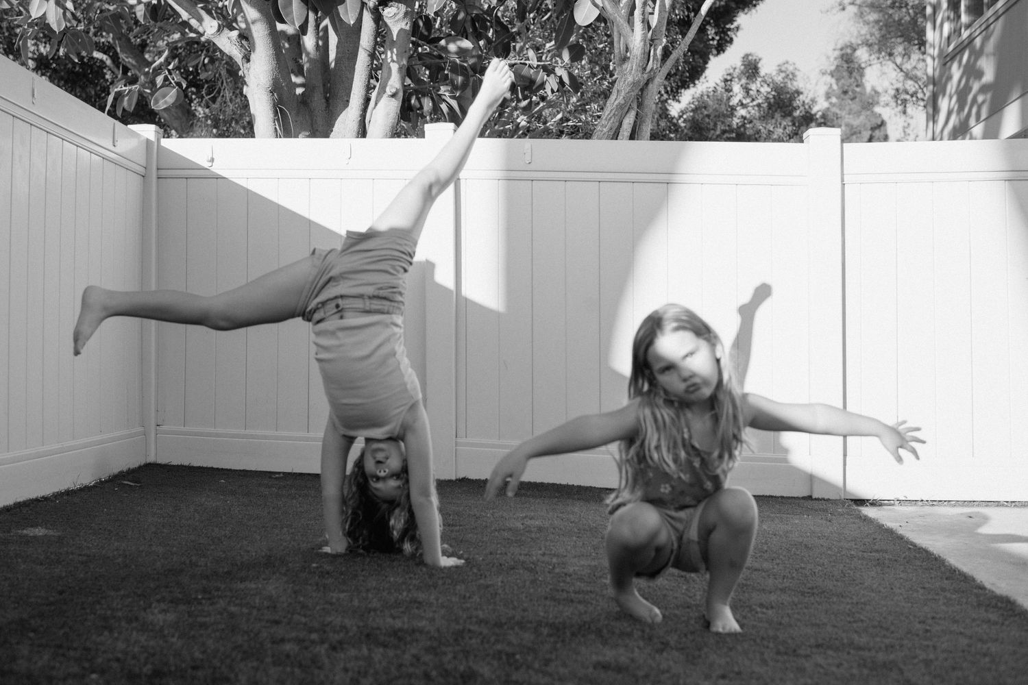 Two girls play in a backyard, one doing a cartwheel while the other poses, in black and white.