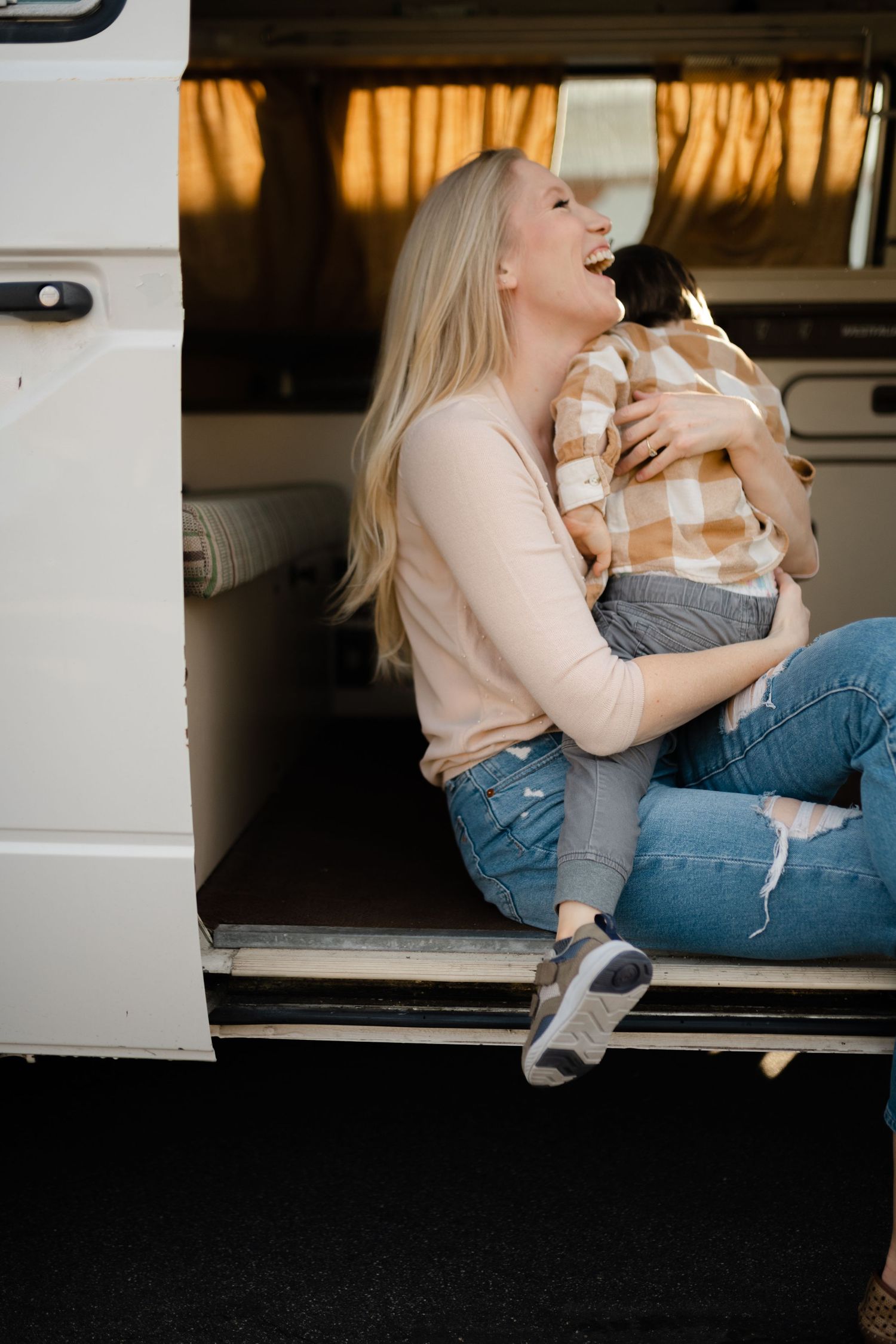 Laughing blonde woman sits in open van doors hugging a young child on her lap.