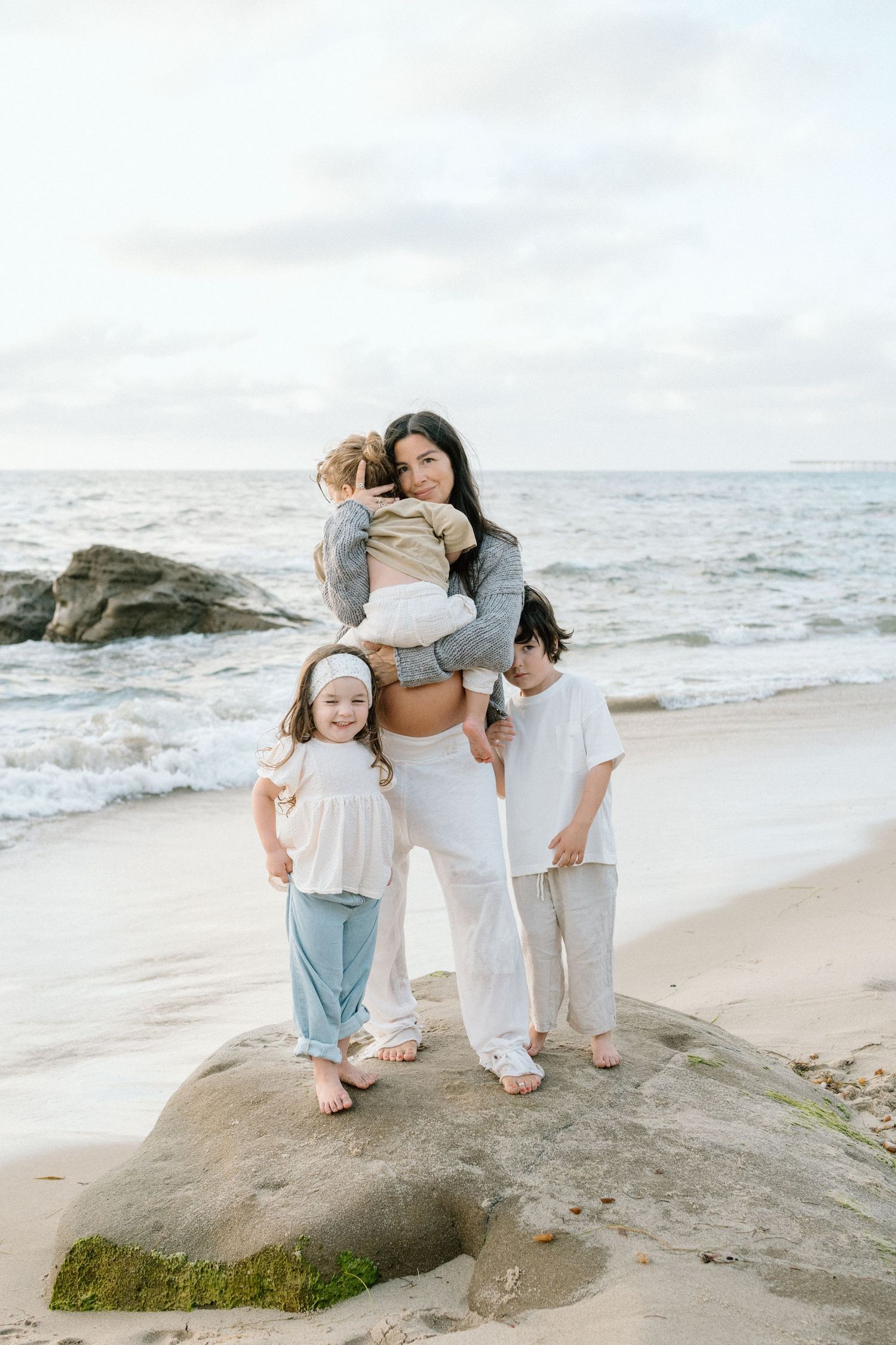A mother holds one child while two others stand on a rock at a sandy beach with ocean waves in the background.