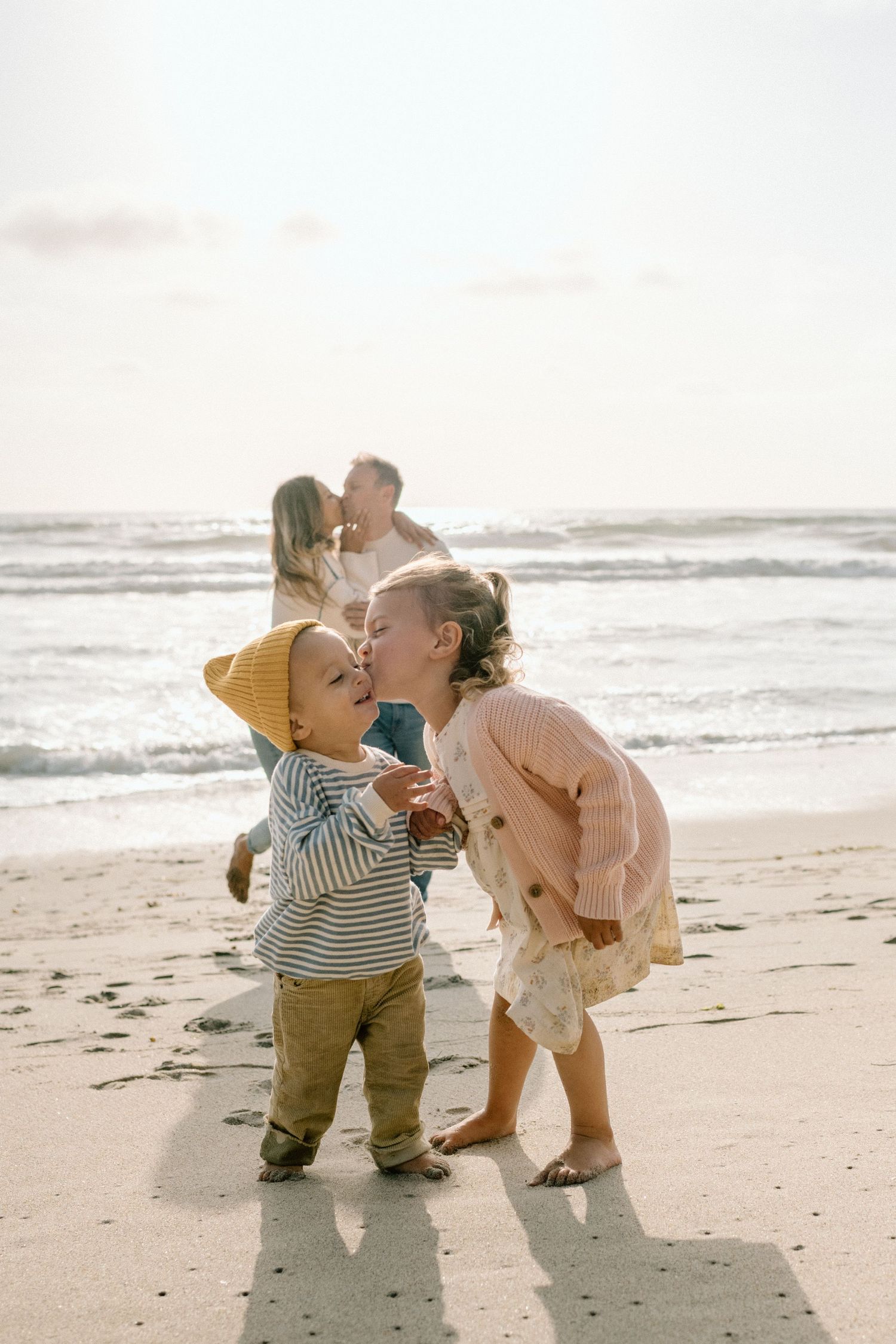 Two young siblings sharing a sweet moment on a sunny beach while their parents embrace in the background near the ocean.