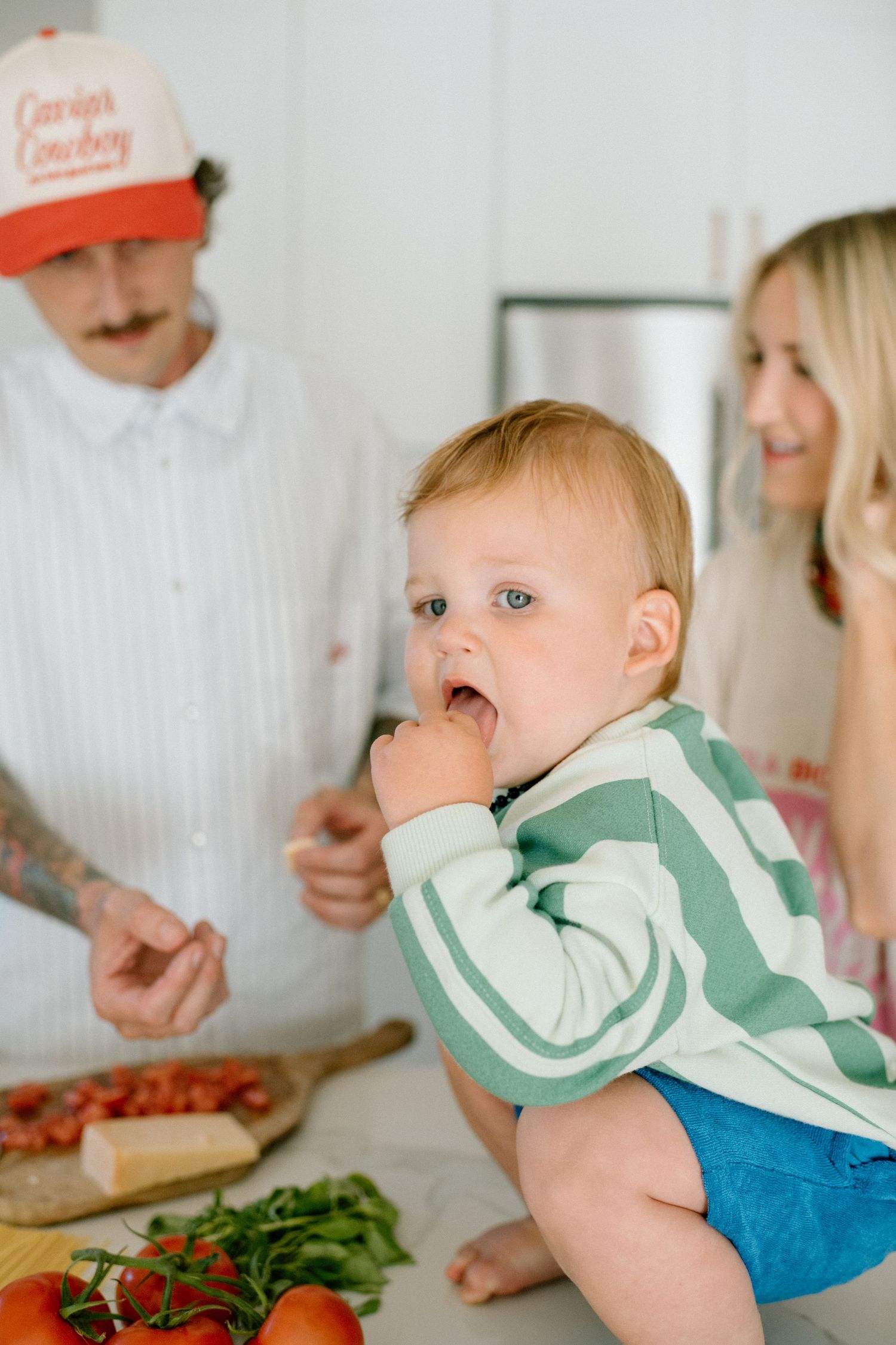 Baby in a green and white sweater watches family preparing food with fresh vegetables on kitchen counter.