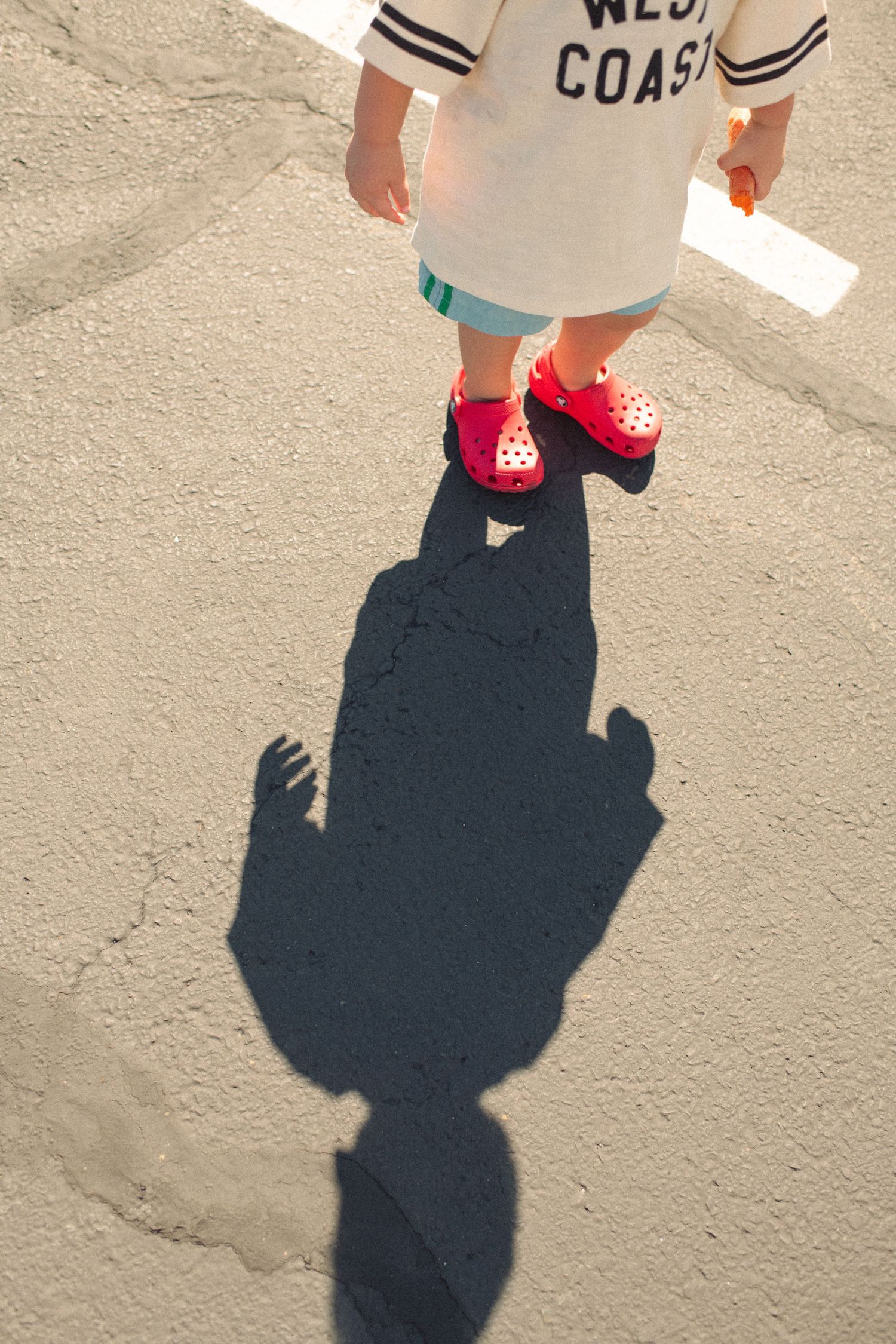 Child in red Crocs and West Coast shirt casting a shadow on pavement.