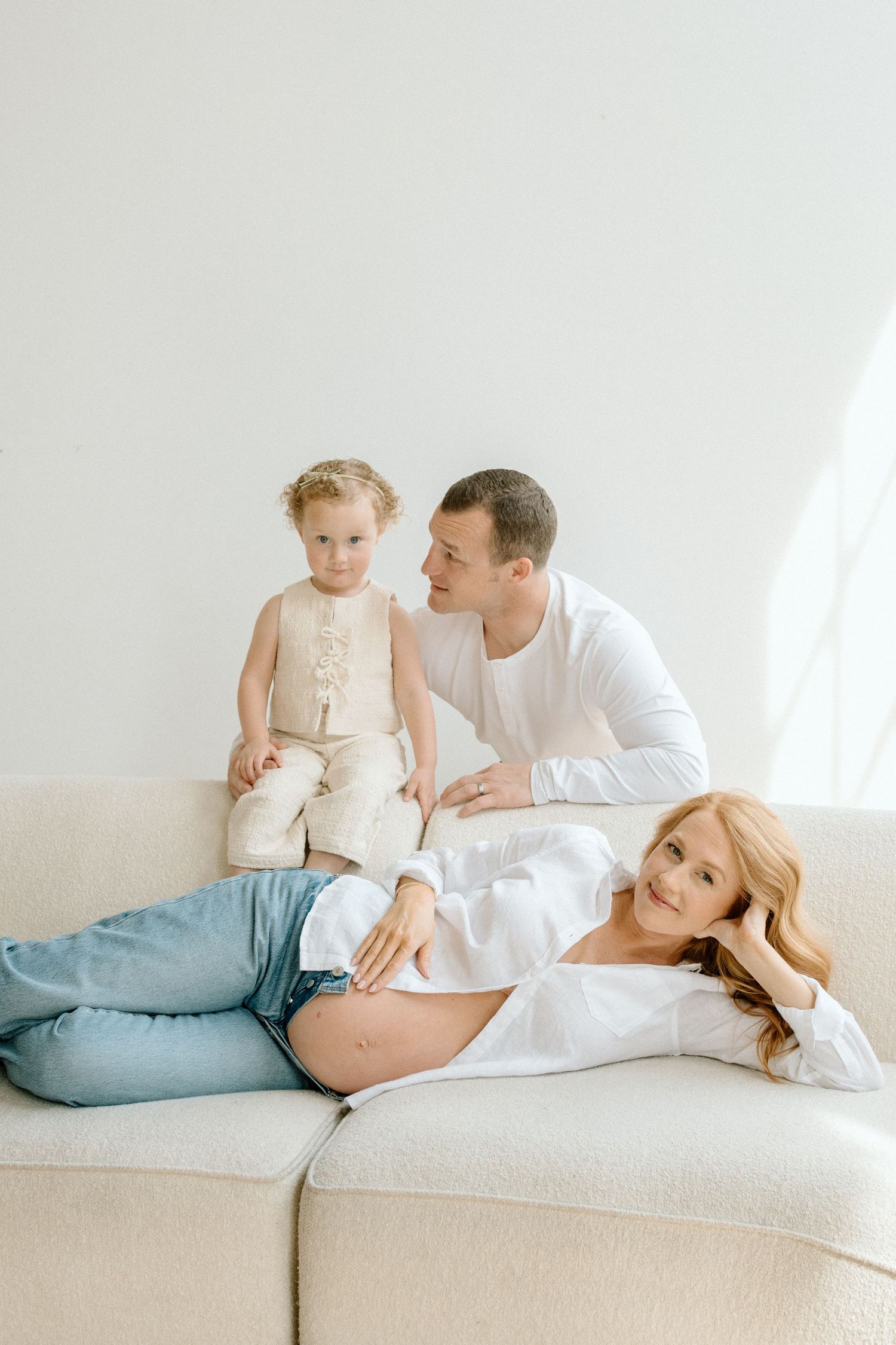 Pregnant woman lying on a sofa with her husband and toddler in a bright room.