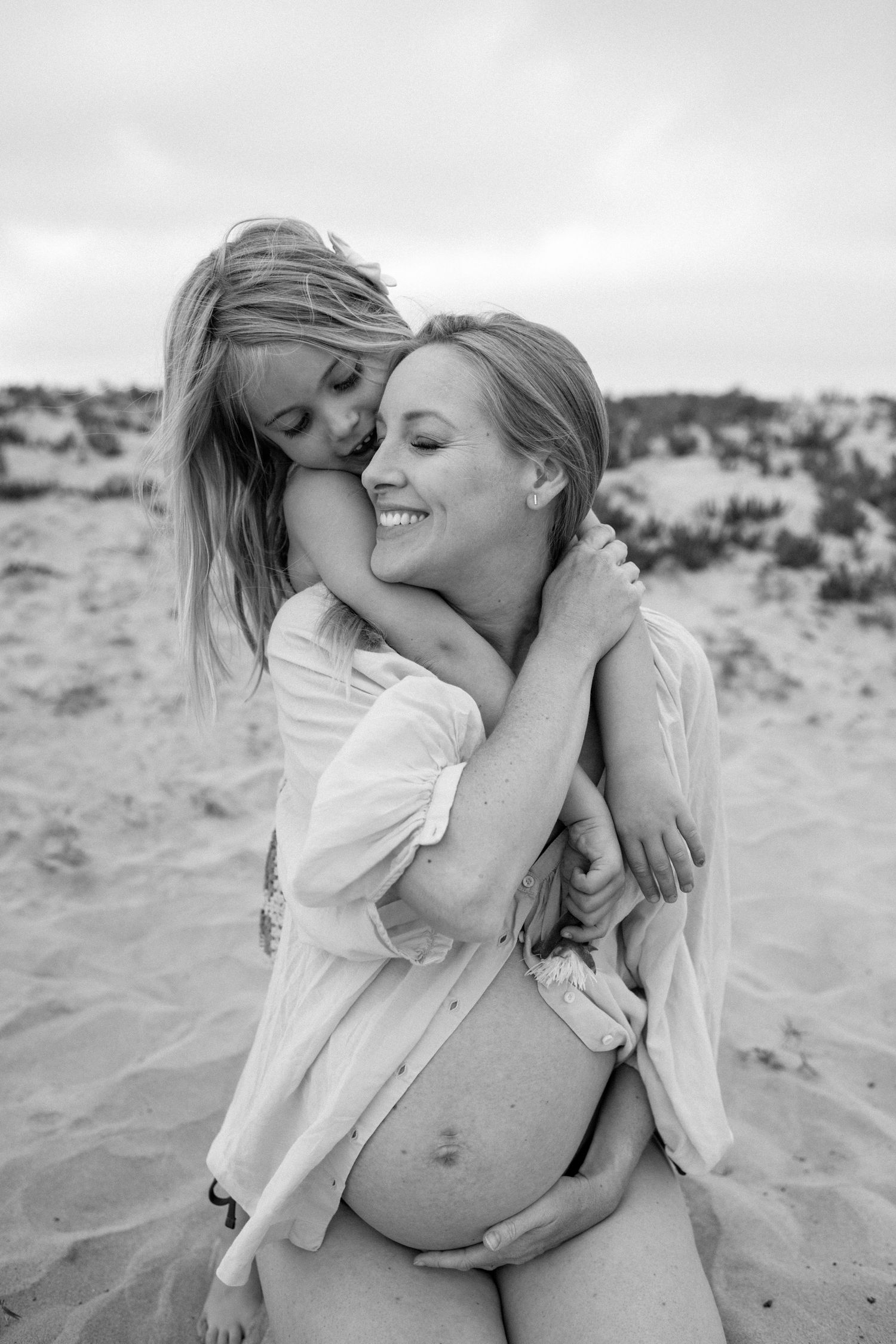 Black and white photo of young girl hugging and kissing smiling pregnant mother on sandy beach.