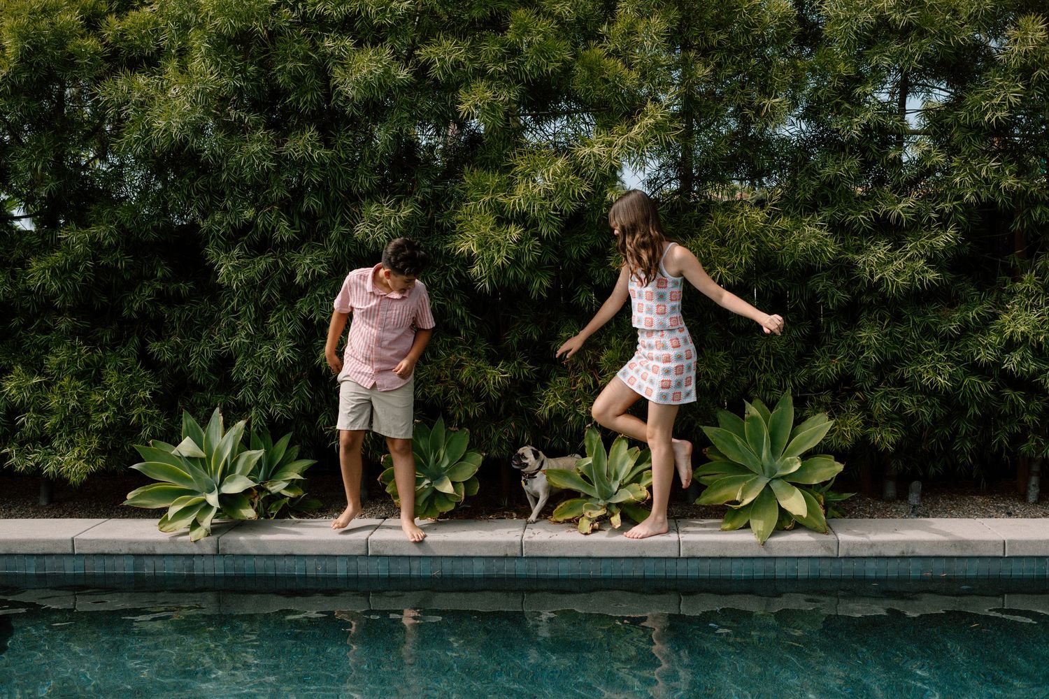 Two kids and a small dog play on the edge of a backyard swimming pool surrounded by lush greenery.